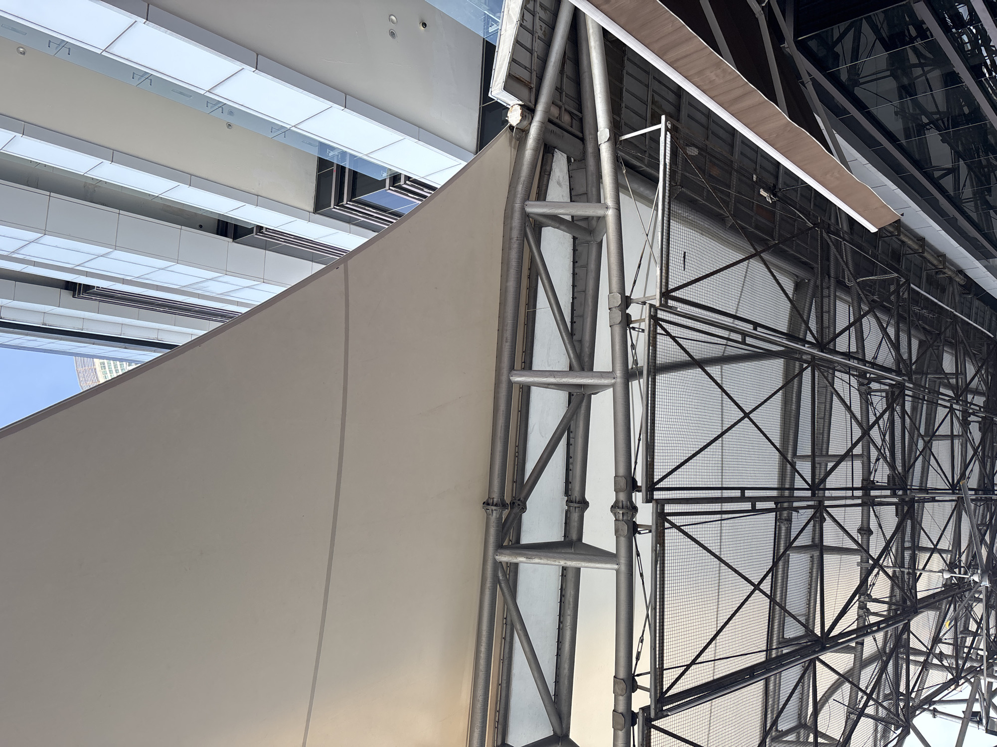 The image shows a low-angle, upward view of a structure in North Jakarta, Indonesia. The primary elements include a large, smooth, off-white wall or panel on the left, constructed from concrete-like material. To the right of the wall, a complex steel structure is visible, constructed of scaffolding, including ladder rungs and cross-bracing. Behind the steel structure, there appears to be a metal mesh or netting. Above the wall, modern architecture can be seen, with a combination of glass and white paneling. The overall impression is an outdoor or semi-outdoor urban environment, with a mix of structural materials.