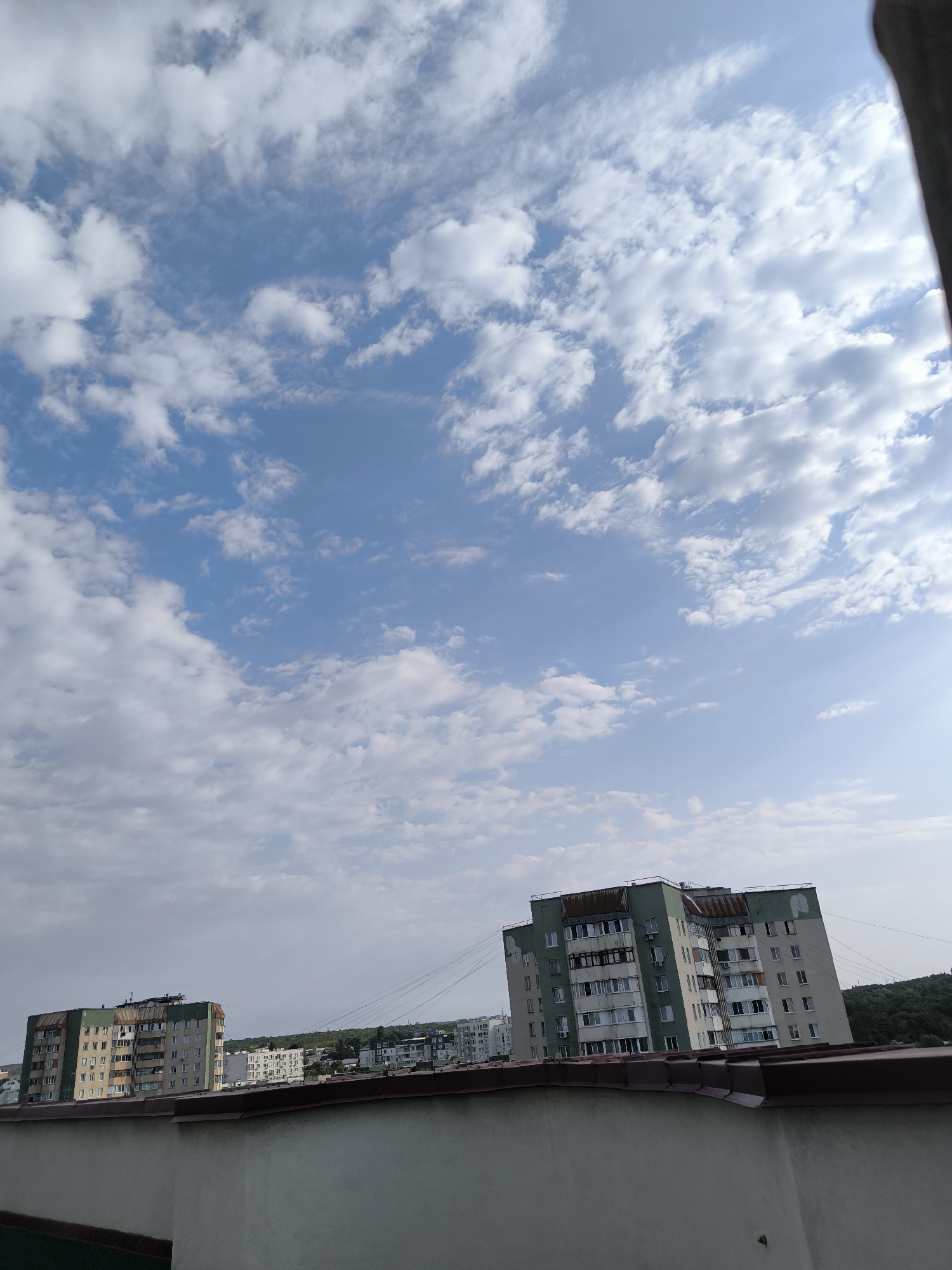 This is a slightly high-angle, full outdoor shot of a cityscape in Chișinău, Moldova, dominated by a bright blue sky with scattered white clouds. In the foreground, a textured light gray wall runs horizontally across the bottom of the frame, topped by a dark reddish-brown, corrugated roofline. Two apartment buildings are visible in the mid-ground. The building on the left is lighter in color with a green roof and appears to be shorter than the one to its right. The building on the right is taller, with a distinct green and light gray facade, and shows signs of wear, including some rust on the roof. Several other buildings of varying heights and architectural styles are visible further in the background, along with a line of utility poles with numerous wires stretching across the scene. A dense cluster of trees can be seen on a hill in the far distance to the right. The lighting suggests it is daytime, likely late morning or early afternoon, with the sun illuminating the sky from the right. There are no people or animals visible in the image.