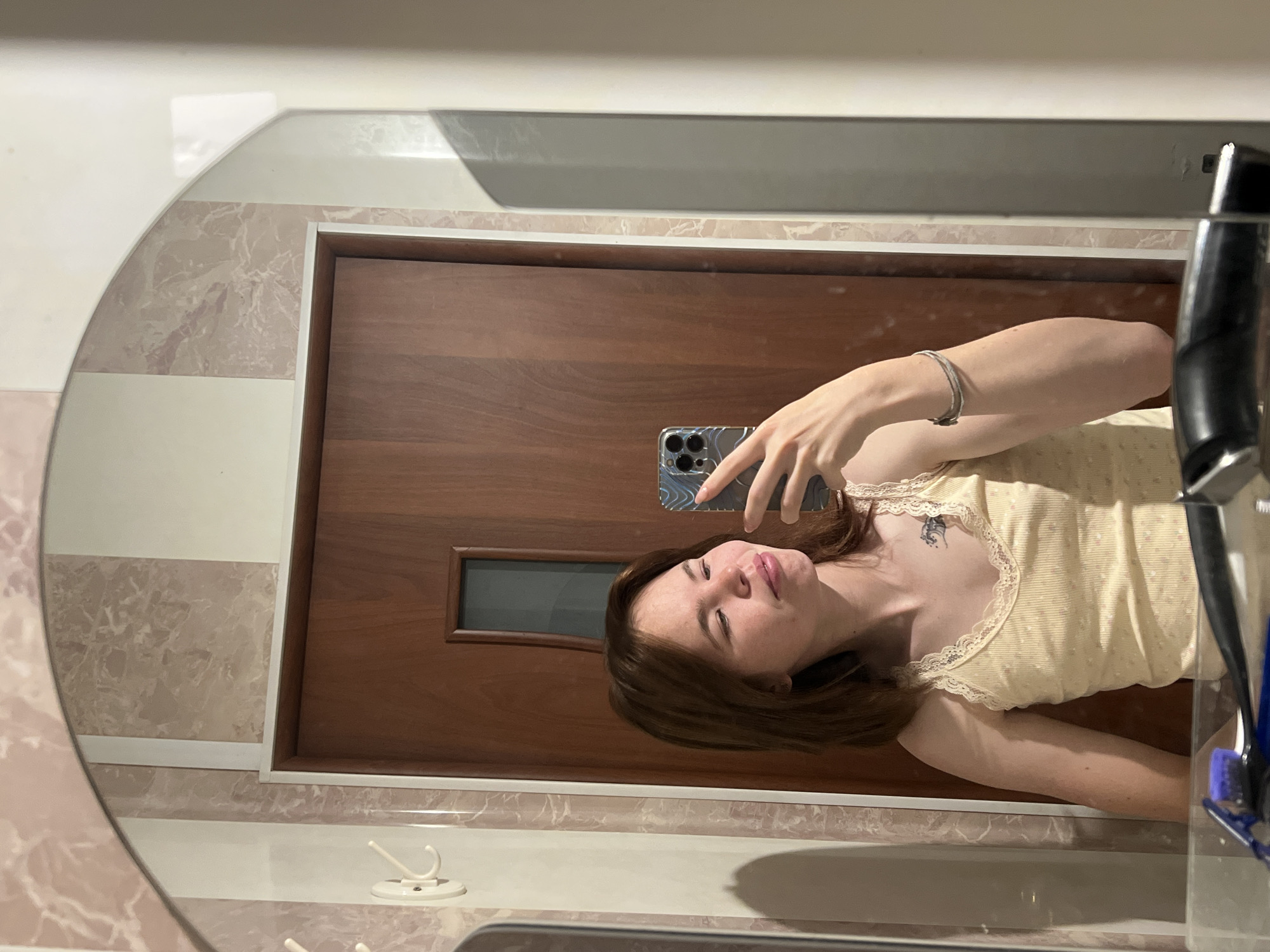 A young woman is depicted in a mirror selfie, with the image rotated 90 degrees clockwise. She is shown from the chest up, wearing a light-colored, possibly yellow or cream, lace-trimmed tank top. A small, dark tattoo is visible on her upper chest/shoulder. Her head is tilted slightly back, eyes partially closed or looking downwards, and lips slightly parted. She holds a smartphone with a patterned back cover in her raised left hand, taking the photograph. A silver-colored bracelet adorns her right wrist.
The oval mirror reflects a background featuring a dark brown wooden door with a small, rectangular, dark glass panel. Surrounding the door and extending into the reflection are tiled surfaces, comprising both plain white sections and light pink/beige marble patterns. Below the door's reflection, on what appears to be a countertop or shelf, a white two-pronged wall hook is visible to the left. To the far right, partially obscured, are some personal hygiene items, including a blue toothbrush. The indoor setting, likely a bathroom or hallway, is illuminated by artificial light, creating soft shadows. There is no visible text.