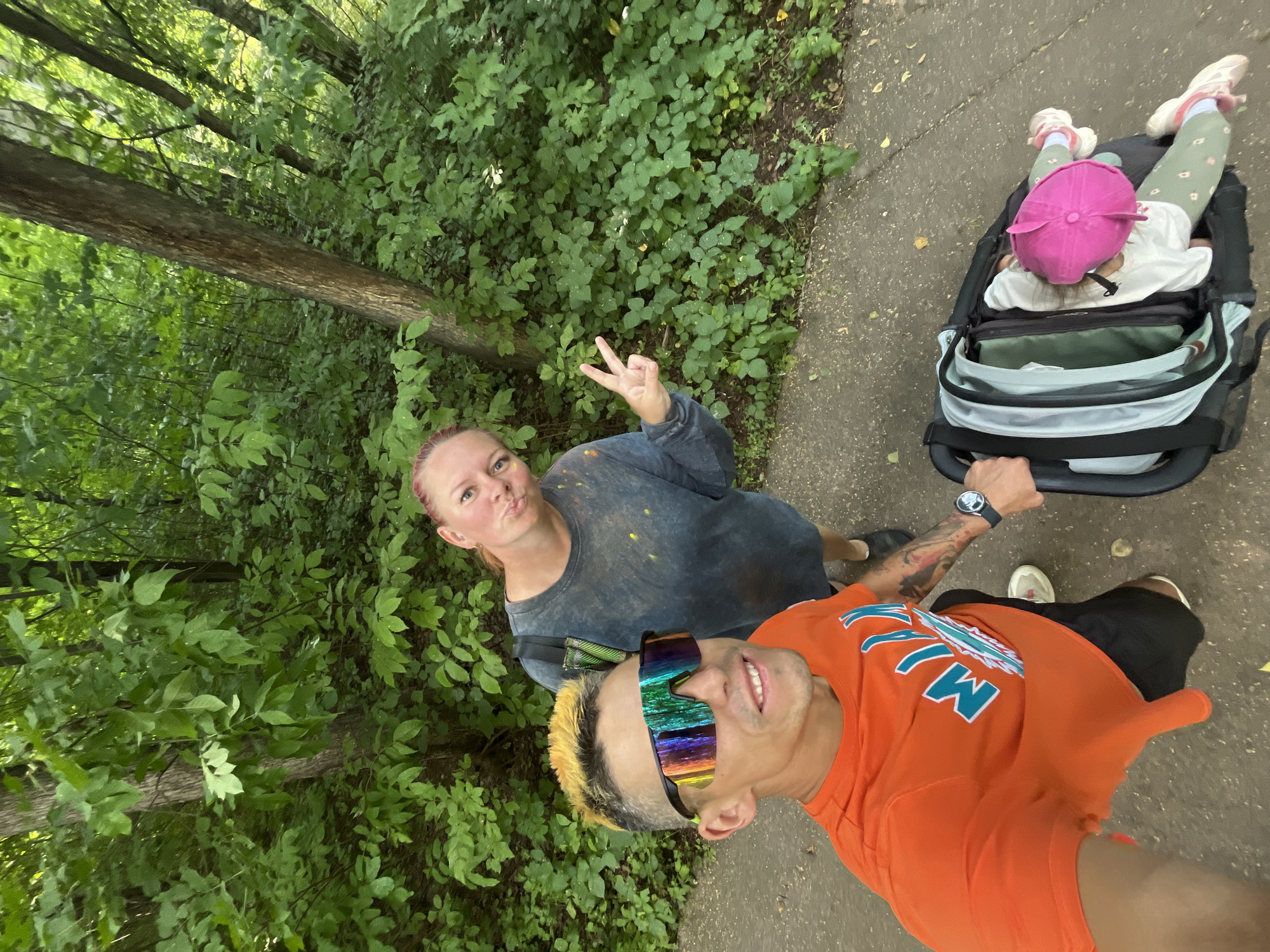 A man, a woman, and a baby are captured in a selfie outdoors. The man, positioned in the foreground, wears an orange t-shirt with a blue and white graphic and black shorts. He has brightly dyed yellow and orange hair and is wearing reflective sunglasses. The woman stands behind him, making a peace sign with her right hand and puckering her lips. She is wearing a dark blue, possibly tie-dyed, long-sleeved shirt, and appears to have colorful paint or dust on her clothing. In a stroller next to the man, a baby wearing a pink hat and a white shirt is visible. The background is a densely wooded area with green foliage and trees, suggesting a park or forest. The ground is a paved pathway. The overall scene suggests a casual outing, possibly a family walk. The specific location is the city of Chaikovsky, Russia.