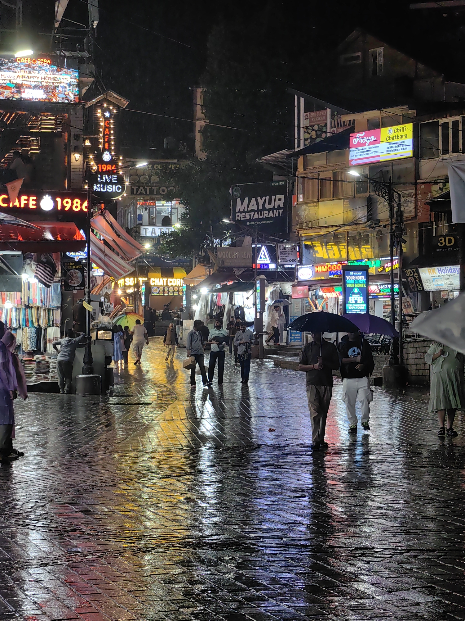 The image depicts a nighttime scene on a wet, cobbled street in Manali, India. The street is lined with shops and businesses, many of which are illuminated by neon signs and lights. The reflections of these lights on the wet pavement create a colorful and vibrant atmosphere. Several people are visible walking on the street, some holding umbrellas to shield themselves from the rain. The architecture suggests a commercial area with multi-story buildings housing various establishments like restaurants, cafes, and shops. The overall impression is that of a bustling street during a rainy evening.