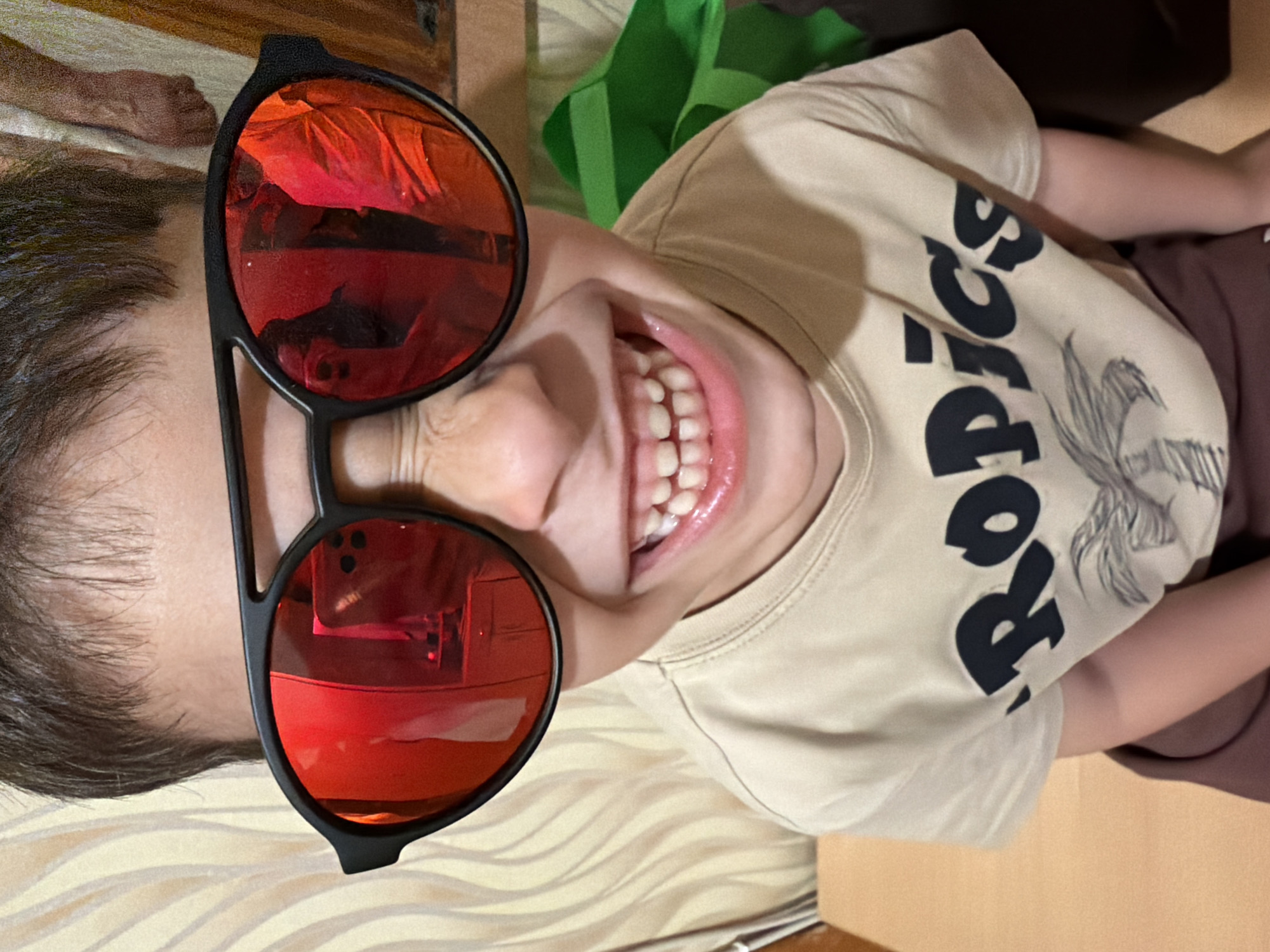 A close-up shot shows a child wearing large, red-tinted sunglasses. The child is smiling widely, revealing a set of white teeth. They are wearing a tan t-shirt with the word "Tropics" printed in large black letters, along with a graphic of a palm tree. The child's head is tilted, and they are looking directly at the camera. The reflection in the sunglasses shows a bright, possibly indoor light source and some crumpled fabric. The background is out of focus but appears to be a bed or couch with a striped pattern. There are also hints of green fabric in the background. The image has a playful and happy feel to it. The lighting suggests it could be daytime, but there are no specific cues to determine the time or weather. The location is unknown.