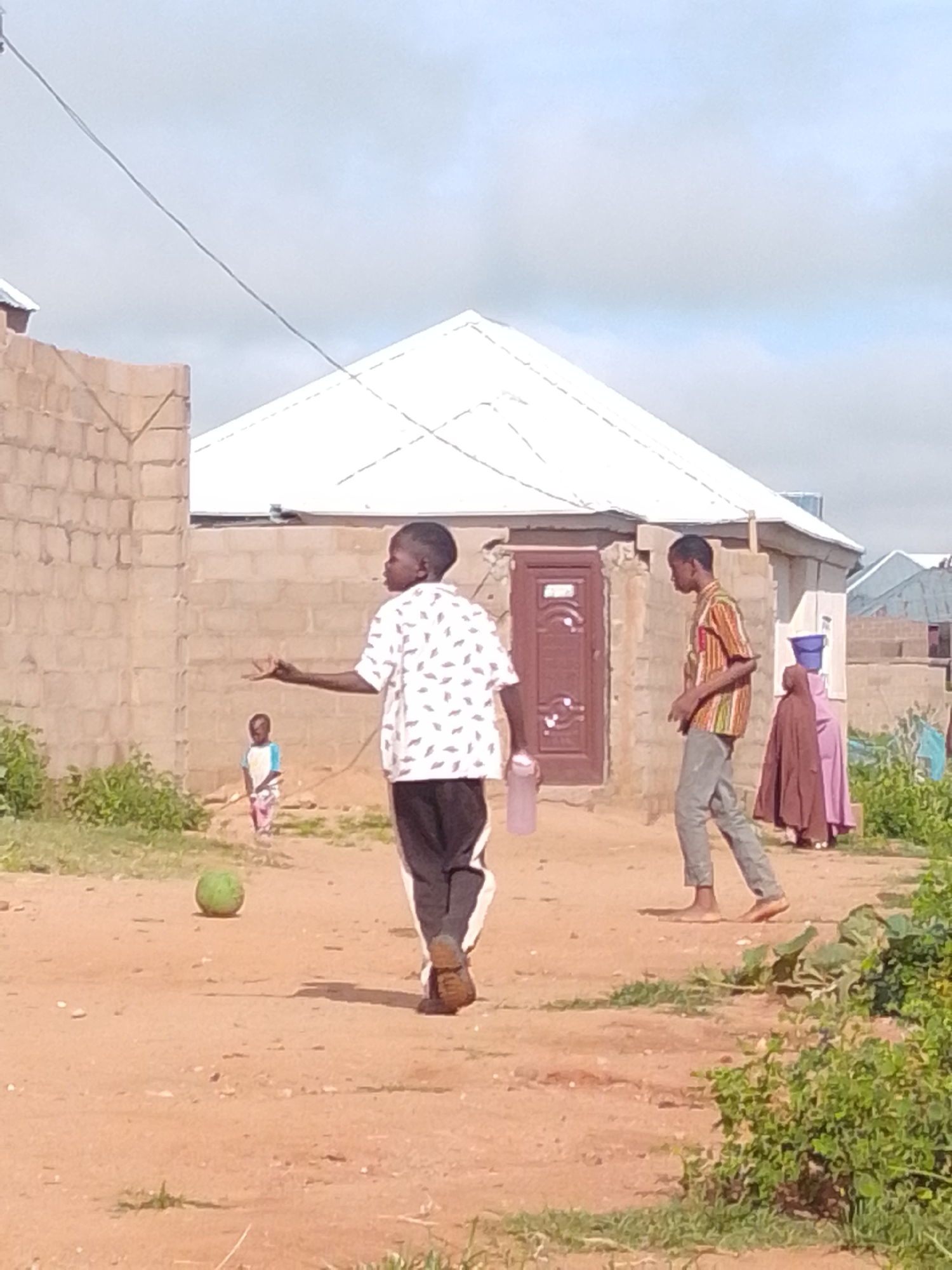 In Tumfure, Nigeria, a group of children are gathered on a dirt path in front of simple, brick structures. A young boy in the foreground, wearing a patterned short-sleeved shirt and dark pants, is captured in mid-stride, facing away from the camera. He holds a pink water bottle in his left hand. To his left and further in the background, another young child stands facing the boy, wearing a light blue and white shirt with colorful striped pants. A young man, dressed in a striped short-sleeved shirt and grey pants, walks past the boy, also facing away from the camera. In the background, two women are partially visible, one in a pink hijab and the other in a darker purple garment. A green, round object, possibly a coconut or a ball, rests on the ground near the foreground boy. The environment suggests a residential area with unfinished brick buildings and overgrown vegetation along the edges of the path. The sky is overcast.