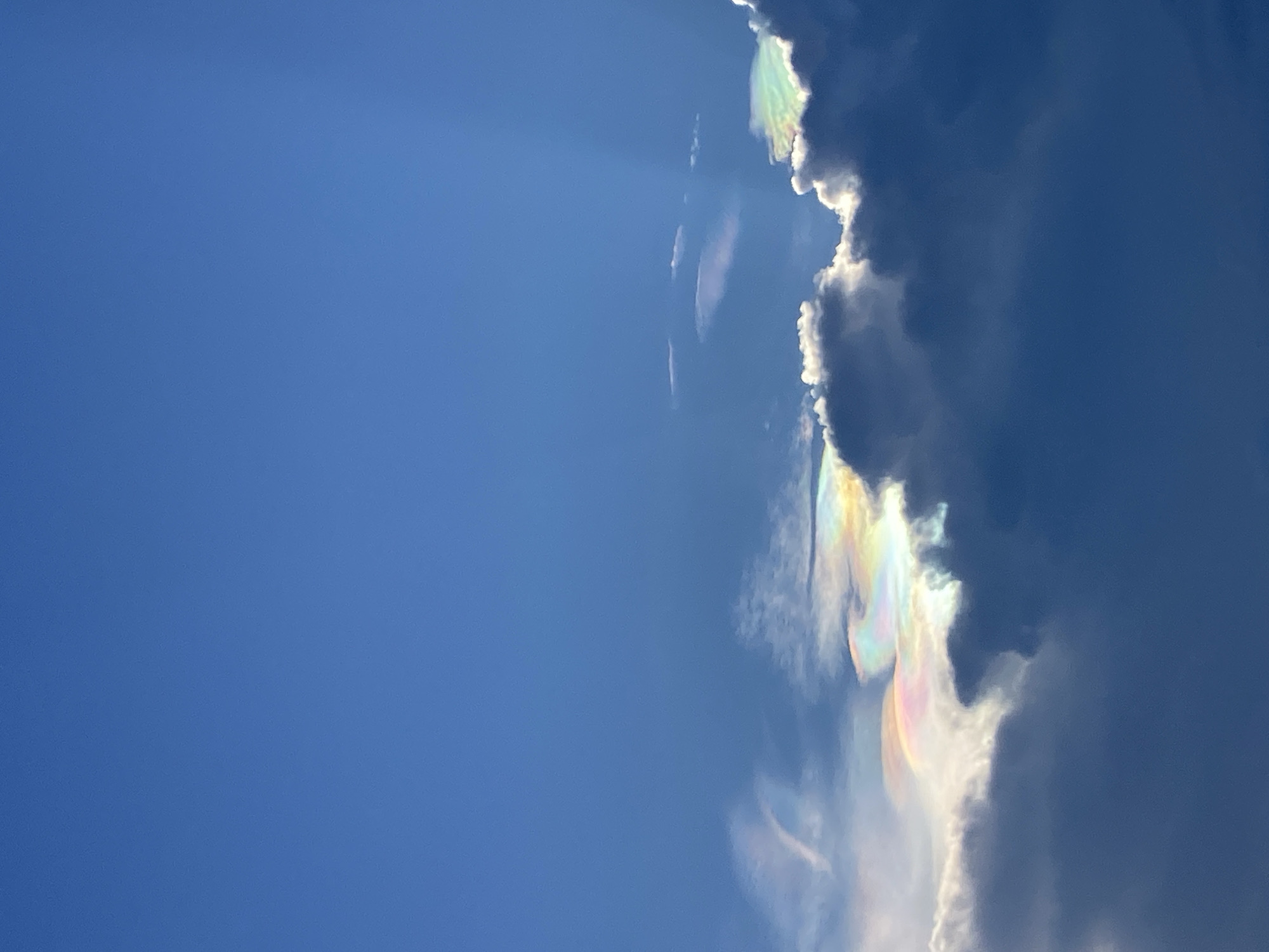 A wide-angle vertical shot captures a vibrant blue sky with scattered clouds, exhibiting iridescent rainbow-like colors. The clouds are illuminated from behind by the sun, causing them to glow with shades of pink, yellow, green, and blue, a phenomenon known as cloud iridescence. The clouds themselves vary in texture and density, with some appearing wispy and thin, while others are more voluminous and dark. The bottom portion of the image is dominated by a large, dark cloud formation, contrasted by the brightly colored, luminous clouds above and to its left. The bright blue sky fills the upper portion of the frame, with faint sunbeams visible on the right side, emanating from an unseen source above. There are no people or man-made objects visible in the frame, focusing solely on the natural atmospheric phenomenon. The scene is consistent with an outdoor environment, likely a sky view from Mũi Né, Vietnam, known for its scenic coastal landscapes and clear skies.
