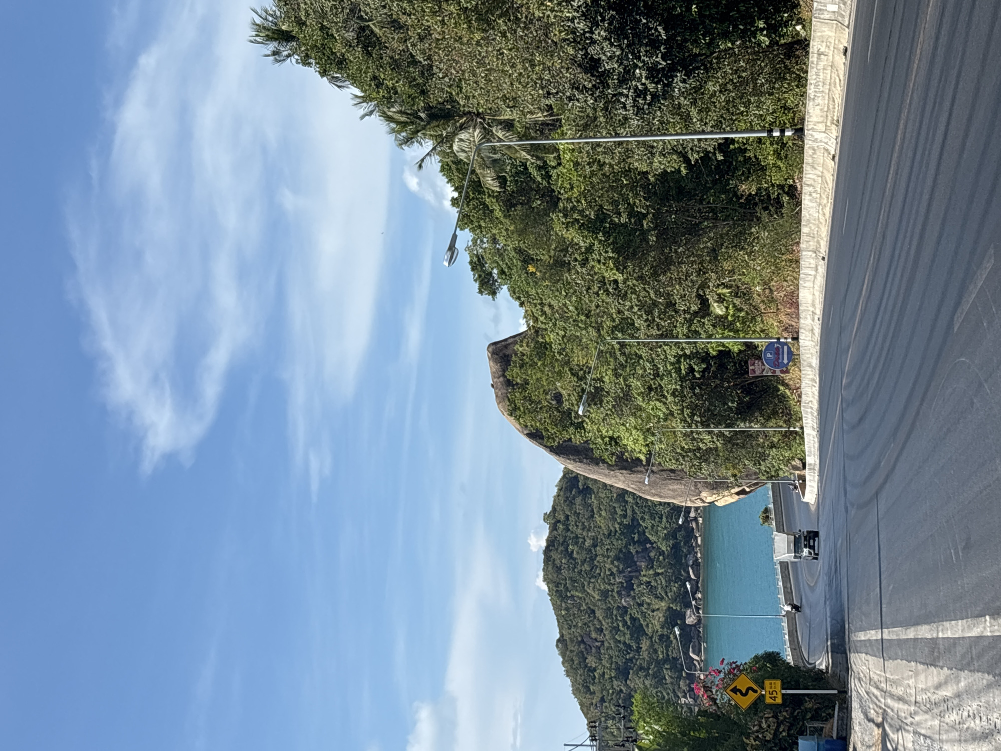 The image shows a road winding along the coast of Baan Chaweng Noi, Thailand. The road curves to the right and is lined with lush green trees and a large rock formation on one side, and the turquoise ocean on the other. Streetlights are positioned along the roadside. A road sign with a yellow background and black markings is visible near the bottom of the image. The sky is blue with scattered white clouds. There is a vehicle traveling along the road.