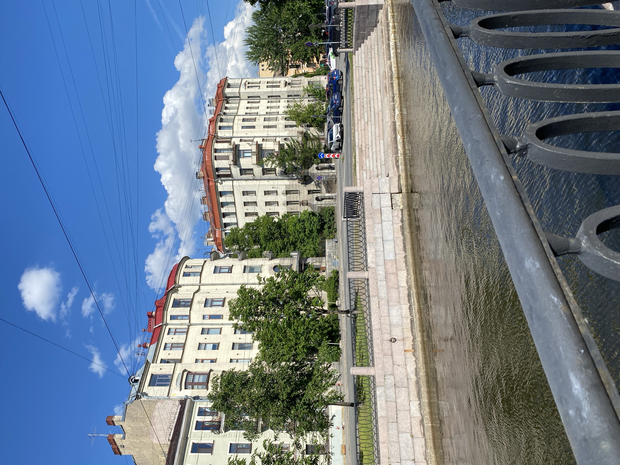 The image captures a vibrant daytime scene along a canal in Saint Petersburg, Russia, likely taken from a bridge. On the far right, a dark metal bridge railing with decorative, circular patterns frames the view.
The central part of the image features a canal with dark, rippling water. A light-colored stone embankment, with a patterned balustrade, runs parallel to the canal on the left bank. Beyond the embankment is a city street where several cars are parked, including blue and white vehicles. Green trees with lush foliage are interspersed along the street, partially obscuring the lower levels of the buildings. A blue and white road sign is visible near the parked cars, though its specific symbols are not discernible.
Further back, a row of multi-story residential or commercial buildings dominates the left side of the frame. These buildings are characterized by their light-colored (cream or beige) facades, numerous windows, and distinctive reddish-orange sloped roofs. Some buildings feature small balconies. The architectural style suggests the historic urban environment typical of Saint Petersburg.
The upper portion of the image reveals a clear, bright blue sky dotted with fluffy white cumulus clouds. Numerous thin, dark power lines stretch across the sky, adding an urban element to the backdrop. The overall impression is one of a sunny day in a picturesque European city. No specific activity or people are prominently featured, presenting a static urban landscape.