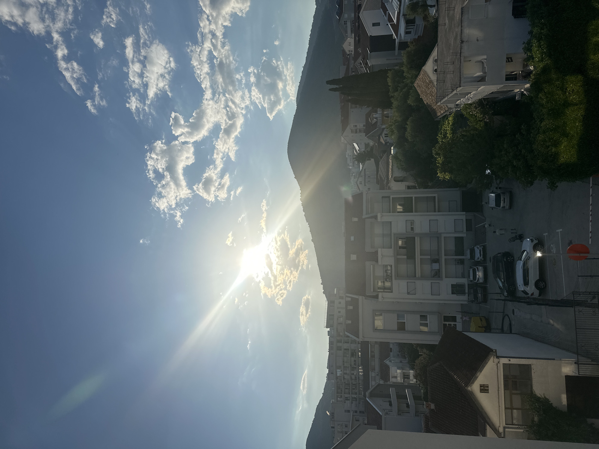 The image depicts a view of the city of Budva, Montenegro, with a mountain range in the background. The sun is shining brightly in the sky, casting rays of light through the clouds. Several buildings, varying in height and architectural style, are visible in the foreground and midground, suggesting a dense urban environment. Some buildings appear to be residential apartments, while others seem to be older, more traditional houses with tiled roofs. Cars are parked in a lot between some of the buildings. Green trees and foliage are interspersed among the structures, indicating that Budva is a city with significant vegetation. The sky is blue with scattered white clouds.