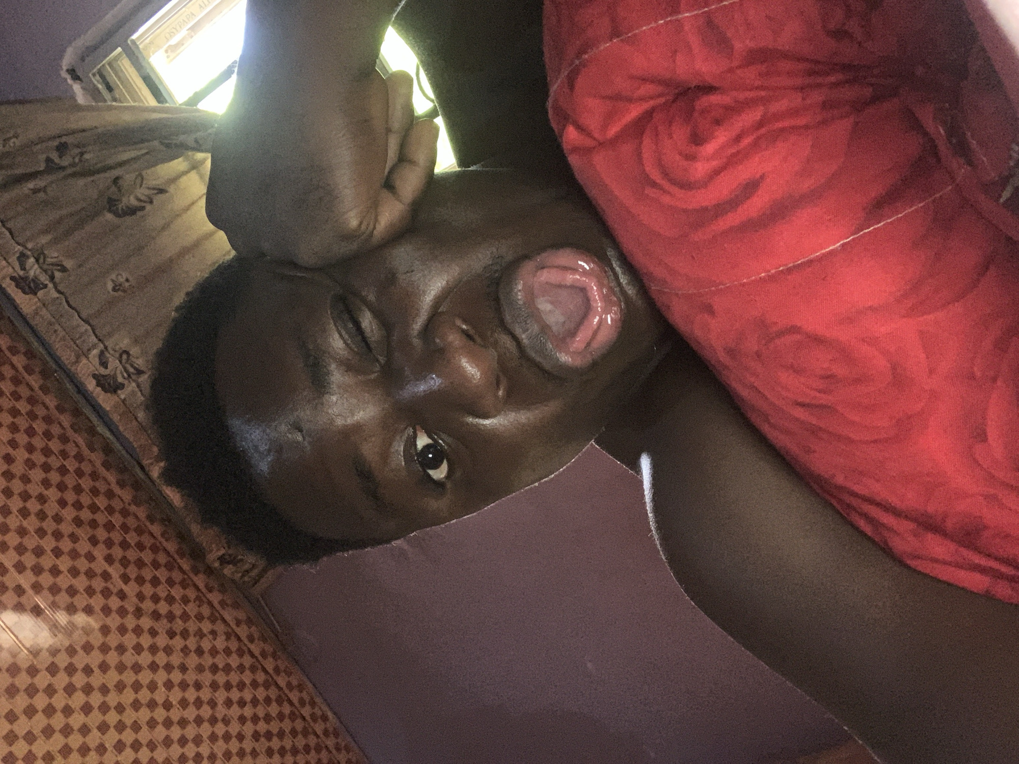 A close-up, slightly distorted perspective shows a dark-skinned person lying on their side, possibly in a room. The person has dark hair and is facing the camera with one eye open and the other closed. Their mouth is slightly open. Part of a red pillow or blanket with a rose pattern is visible to the right of the person's face. The person's hand rests on their forehead.
A brown and tan patterned floor is visible in the lower left corner of the image. A curtained window is visible in the top left of the image.
The location of the photo in Akwanga, Nigeria cannot be determined from the visual information.