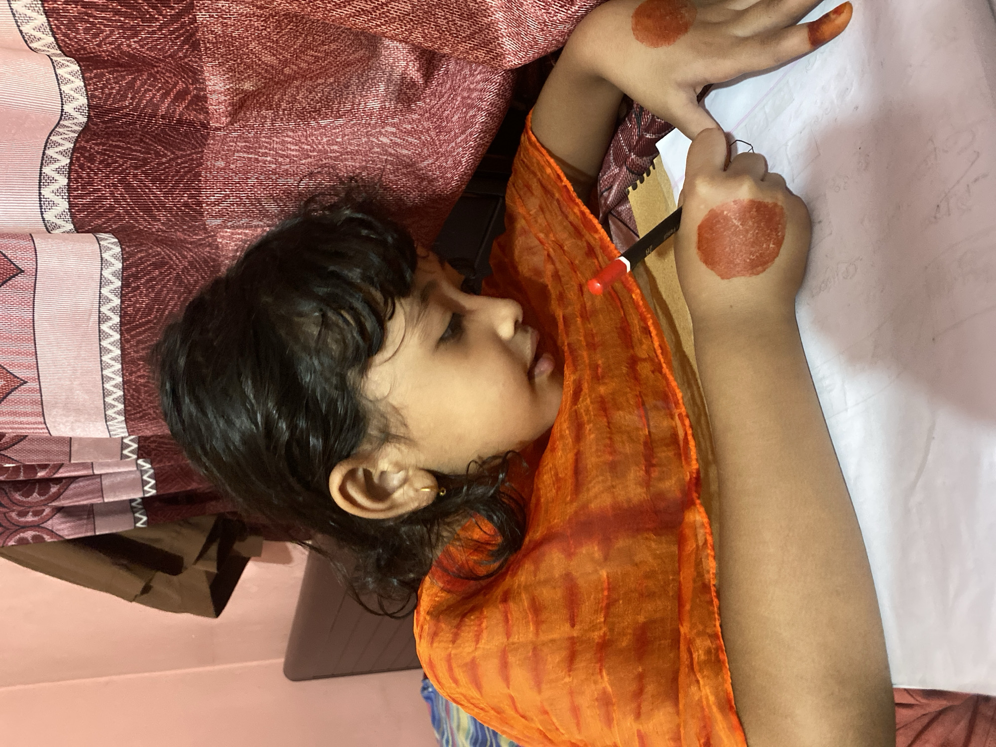 A young child, likely a girl, is captured lying down and intently writing or drawing on a piece of white paper with a pen. The image is oriented sideways, with the child's head to the left. She has dark, wavy hair and wears a vibrant orange, patterned scarf or dupatta. A small golden hoop earring is visible in her right ear.
Both of the child's hands are decorated with circular reddish-brown henna (mehndi) designs on the back, and her fingertips are also stained with henna. She holds a pen, which has a red cap and a black barrel with "Pebeo" and "2B" faintly visible, and is focused on the paper. The white paper shows faint light gray markings, possibly lines or previous writing.
The child is resting on patterned textiles. Behind her, a maroon or reddish fabric with a detailed, repeating pattern is visible, alongside a lighter pink or beige fabric featuring striped and zigzag designs. A dark object, possibly part of furniture or a bag, is partially visible beneath the fabrics near her head. The background consists of a plain, light pink wall. The setting appears to be indoors, consistent with a home environment in Dhaka, Bangladesh, given the cultural elements like the henna and attire. The child's expression shows concentration and engagement in her activity.