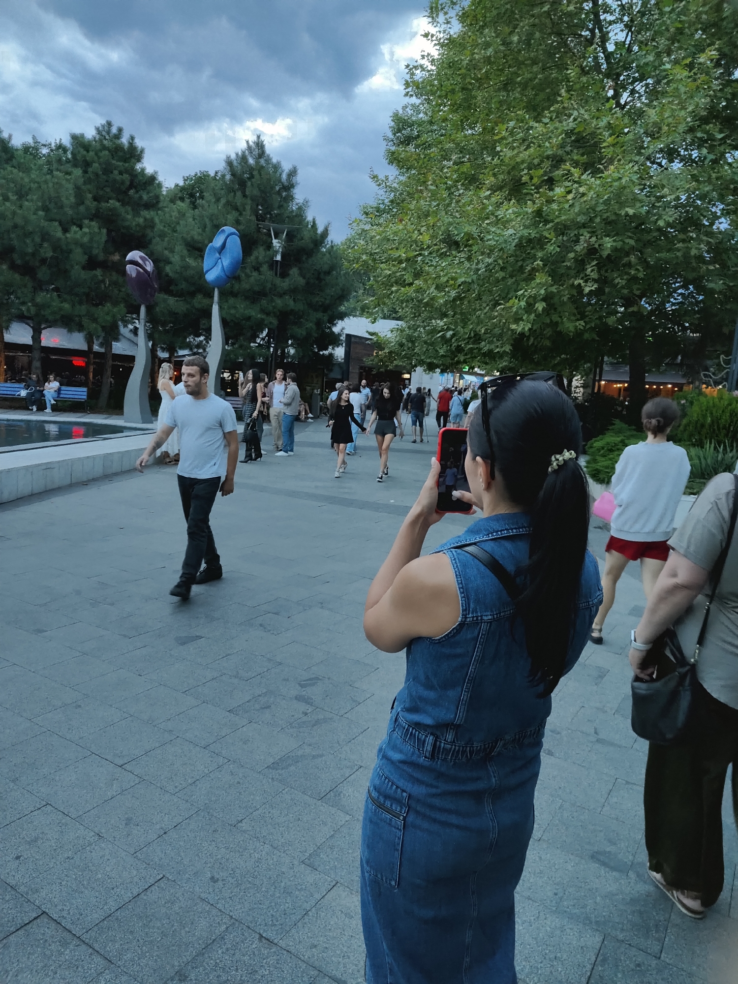 The image depicts an outdoor public space, likely a pedestrian area in the city of Odesa, Ukraine, given the context provided. In the foreground, a woman with dark hair tied in a ponytail, wearing a denim jumpsuit, is holding a red smartphone and taking a picture. She is positioned in the lower right quadrant of the frame, facing away from the camera. To her right, partially visible, is another person wearing a light-colored shirt and dark trousers.
In the mid-ground, a man wearing a light blue t-shirt and dark trousers is walking across the paved area. Several other people are scattered throughout the mid-ground and background, some walking and others sitting on benches. The environment includes trees with green foliage, decorative sculptures resembling large bean-like objects in purple and blue, and buildings in the background. The ground is paved with large rectangular grey tiles. The sky above is overcast with grey clouds.
