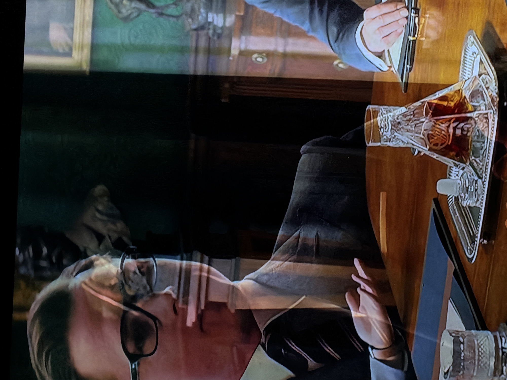 The image captures a reflected view of a person watching a screen, showing a scene in what appears to be a dimly lit, upscale study or office. The original image is rotated 90 degrees clockwise; this description assumes a correct upright orientation.
**Subjects:**
The primary subject in reflection is a person with short, light-colored hair, wearing black-rimmed glasses, looking towards the right side of the frame. Their left hand, with fingers crossed in a gesture often associated with hope or good luck, is also visible in reflection in the lower-right quadrant.
The scene on the screen features a man's arm and hand holding a black and gold fountain pen in the upper right. He is wearing a blue-grey suit jacket over a white shirt with cufflinks. To the left, part of another person's arm or hand is vaguely visible near a framed painting.
**Objects:**
A prominent cut-glass decanter, filled with an amber liquid (likely whiskey or brandy), rests on a decorative silver or chrome tray on a polished wooden table. The table appears to be a desk, made of rich, dark wood with intricate details. A framed painting with an indistinct dark figure hangs on the wall in the background to the left. Dark, traditional furniture is suggested in the room's background.
**Scene/Setting:**
The on-screen scene depicts an interior setting, possibly a classic study or office, characterized by the wooden desk, decanter, and painting, suggesting a formal or contemplative atmosphere. The reflections suggest the viewer is in a darker environment, likely watching the screen in a room with some ambient light.
**Activity or Event:**
On the screen, the man is holding a pen, poised as if to write or having just written. The scene evokes a moment of thought or decision. The reflected person is observing this scene; their crossed fingers might imply their engagement or hope related to the on-screen narrative.
**Notable Details:**
The image is dominated by the strong, semi-transparent reflections of the viewer and their hand over the depicted scene. The lighting within the on-screen scene appears to be artificial and warm, highlighting the polished wood and the decanter's contents. There is no visible text.
**Location Context:** Sofiivska Borshchahivka, Ukraine.