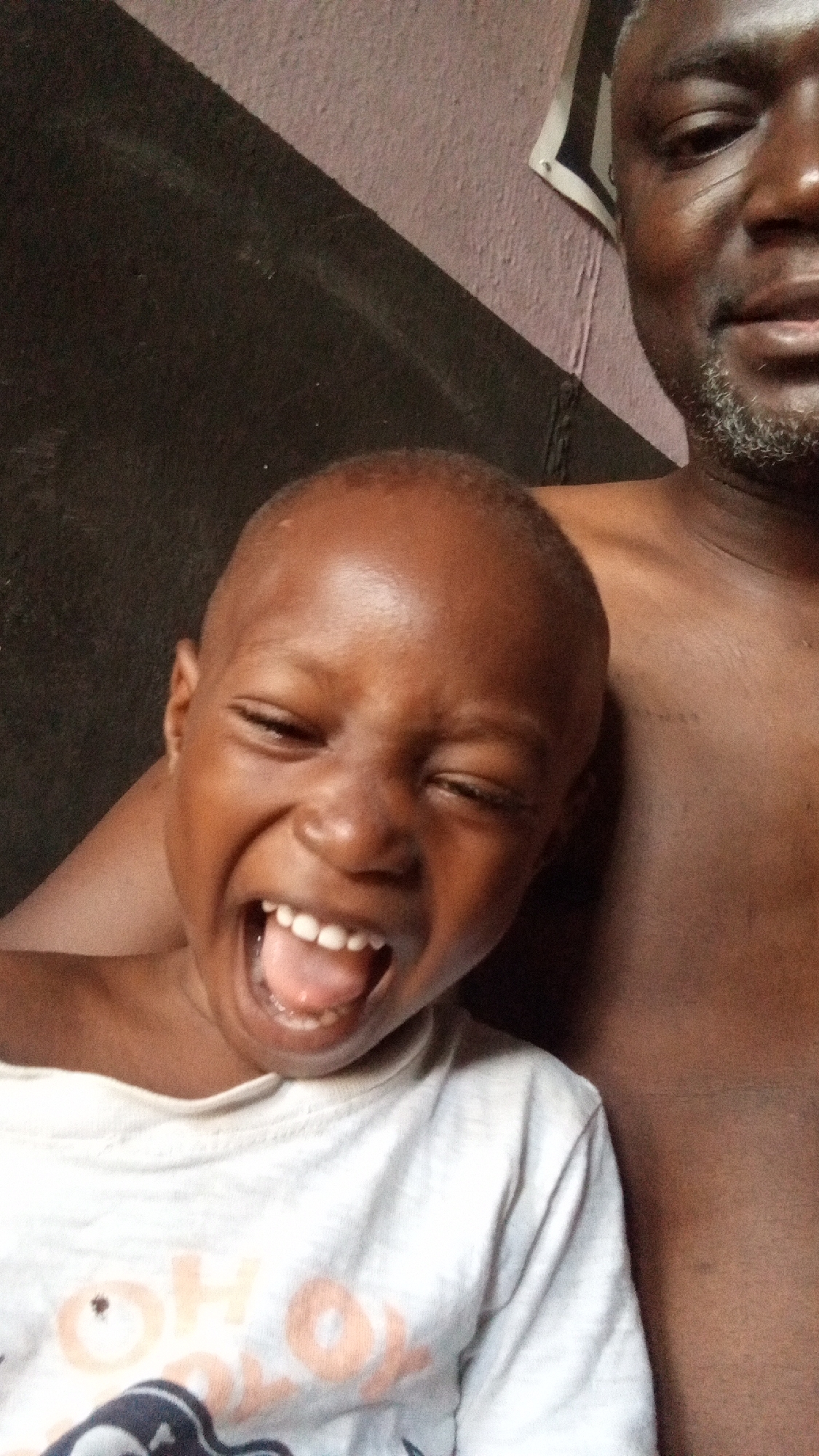 A close-up, informal portrait features a laughing child and a partially visible adult, likely in an indoor setting in Sabon Tasha, Nigeria.
The prominent subject is a young child, appearing to be a boy, with dark skin and a shaved head. He is captured mid-laugh or shout, with his eyes squinted shut and his mouth wide open, revealing several white teeth and a pink tongue. He is wearing a light-colored, long-sleeved t-shirt, possibly white, with some creases. Faint orange lettering is visible on the t-shirt, which appears to spell "OHO" followed by additional, indistinct letters that might form words like "HOX" or "BOX".
To the right and slightly behind the child is an adult male, with only his right shoulder, part of his chest, and the right side of his face visible. He has dark skin, short dark hair, and a salt-and-pepper beard. He appears to be shirtless. The child's head is resting near the adult's shoulder, suggesting a close relationship, likely parent and child.
The background consists of a wall with two distinct color sections: a dark, possibly black or very dark brown area in the lower left, and a lighter, faded purplish or off-white section in the upper right, separated by a diagonal line. A small piece of paper or a poster with some indistinguishable print is tacked to the lighter wall section behind the adult's head. The child's expression conveys uninhibited joy and happiness. The lighting suggests an indoor setting during the day.