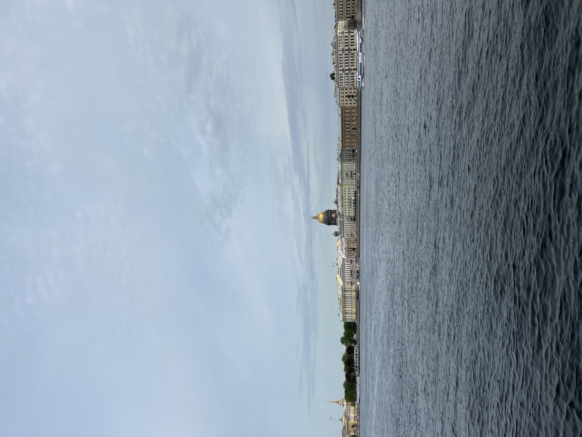 The image captures a panoramic view of Saint Petersburg, Russia, from across the Neva River. The sky is overcast, with a pale blue hue punctuated by streaks of clouds. The river's surface is dark, with visible ripples suggesting movement.
The city skyline is dominated by several prominent landmarks. A golden dome, likely belonging to Saint Isaac's Cathedral, stands out. Other notable buildings include the Admiralty building, identified by its spire. Various other buildings line the riverfront, showcasing the architectural diversity of the city. A boat is moving down the river.
The overall scene depicts a serene urban landscape with the river acting as a primary feature. The photo is taken in vertical orientation.
