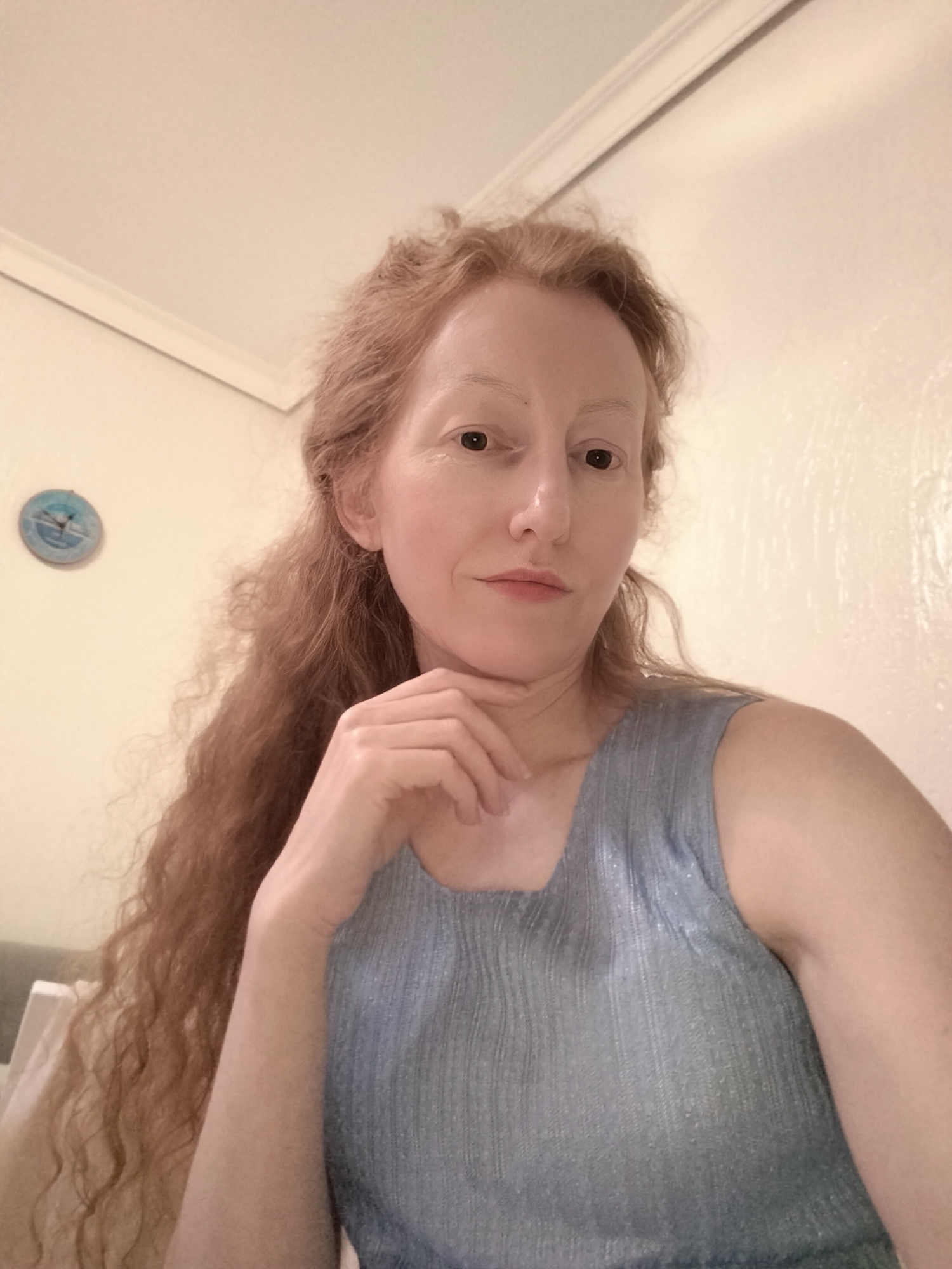 A fair-skinned woman with long, reddish-brown curly hair poses for a selfie indoors. She rests her chin on her left hand. She is wearing a sleeveless, blue-gray textured top. Behind her is a plain white wall with a small, round blue clock. The location is not identifiable as Krykhivtsi, Ukraine from the given image.