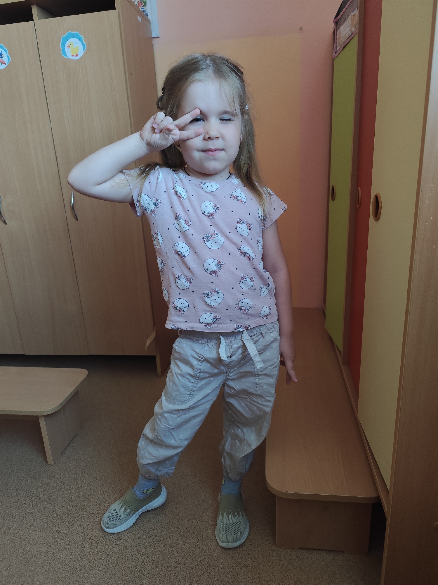 A young girl stands in a room. She is wearing a pink t-shirt with cat faces on it, light-colored pants, and green sneakers with gray socks. She is making a peace sign over her left eye, which is closed. The background consists of wooden cabinets in light brown and yellow/green colors, and a wooden bench. The location is Фоки, Russia.
