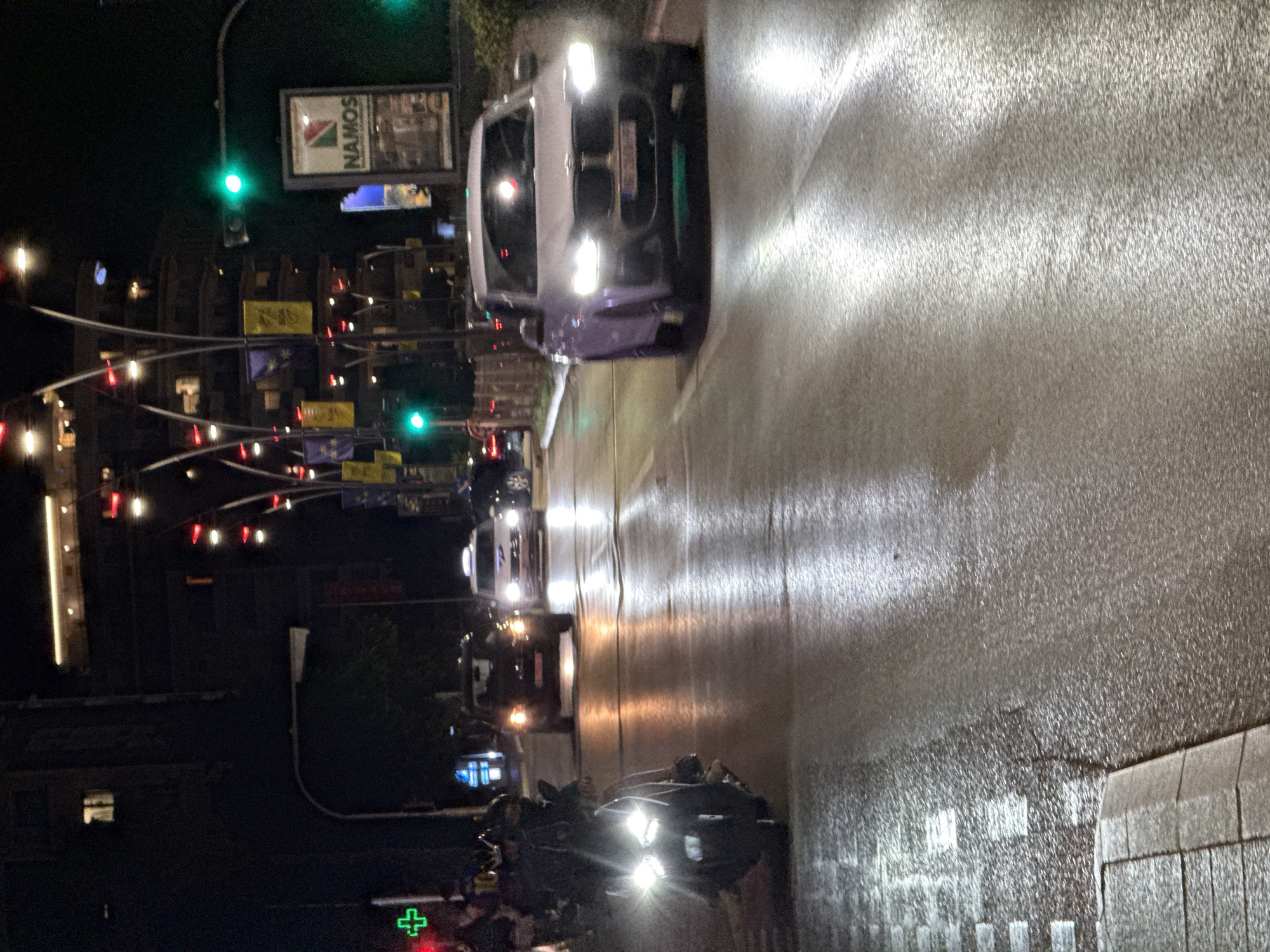 The image depicts a street scene at night in Budva, Montenegro, likely near the coast given the presence of a "NAMOS" billboard which is a well-known establishment in that area. A light purple BMW SUV is in the foreground, moving forward with its headlights on. To the left, a dark motorcycle with its headlights illuminated is also moving forward, with the rider blurred due to motion. Further down the street, a dark sedan and a white taxi are visible, also in motion. The street is wet and reflects the lights from vehicles and streetlights. Buildings with illuminated windows are present in the background. Traffic lights are visible, with at least one showing green. A green cross symbol is also visible on a building to the left. The overall impression is of a busy urban street at night.