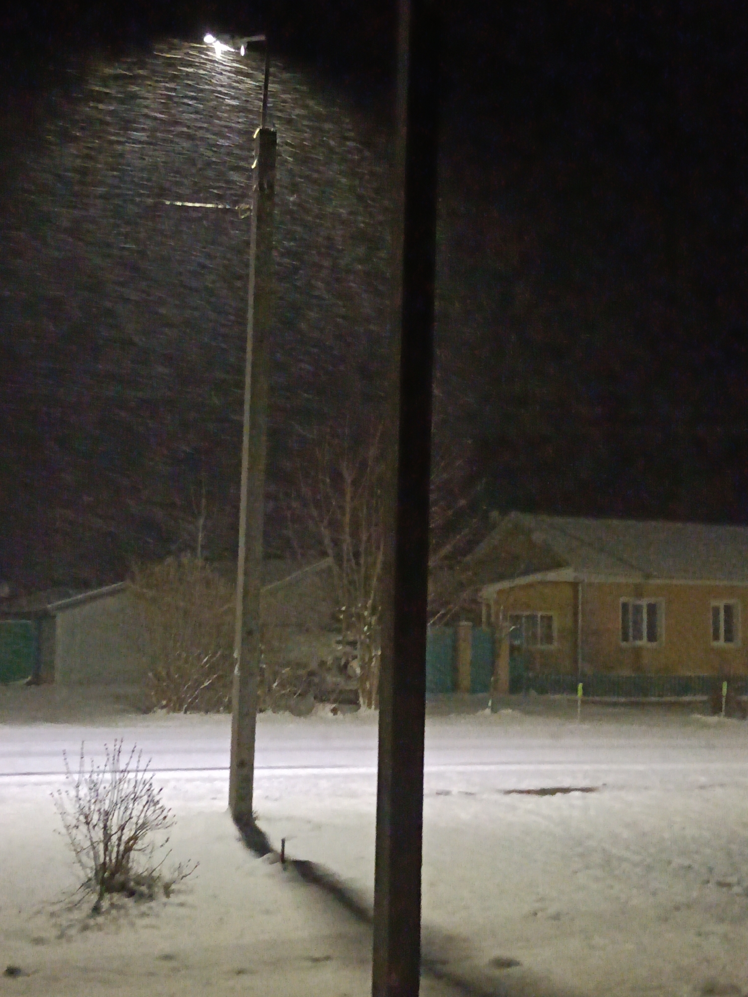 The image depicts a snowy night scene in Красный Маяк, Russia. A streetlight illuminates falling snow near a wooden utility pole. A snow-covered street and yard are visible, with a small, leafless bush in the foreground. A one-story house with multiple windows is in the background, along with a smaller building. A dark vertical object, possibly another pole, is in the lower right of the frame.
