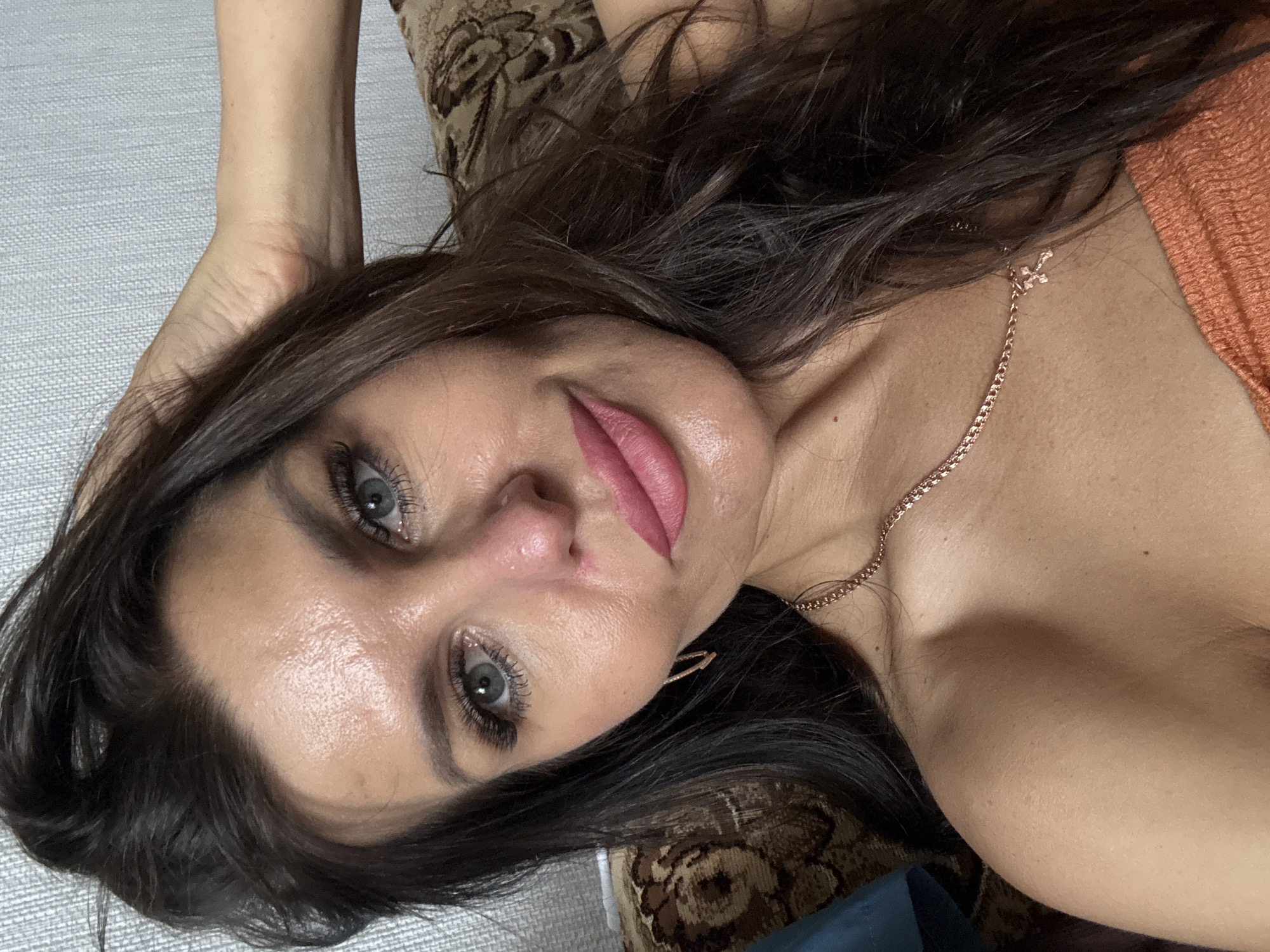 A close-up, eye-level, indoor selfie shows a woman with dark brown hair and light blue eyes, wearing a coral-colored top and a rose gold necklace with a cross pendant. Her right hand is touching her head, and she is looking directly at the camera with a slight smile. The background consists of a patterned sofa. The image quality is good, with a shallow depth of field that blurs the background. There are no visible markings or elements that would indicate the specific location within Dubovaya, Russia.