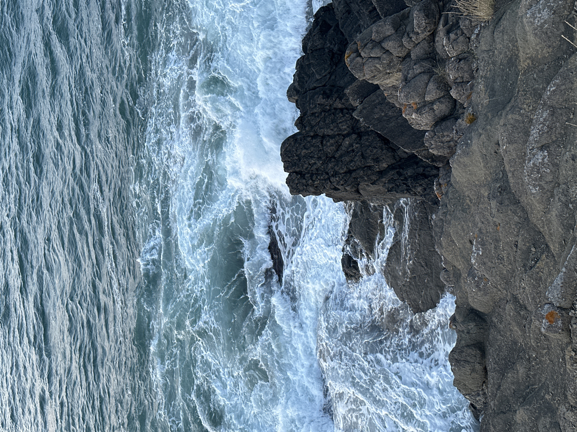 The image depicts a rough sea crashing against a rocky shoreline in Sinemorets, Bulgaria. The water is a deep blue-green with white foam churning around dark, jagged rocks. The foreground is dominated by a grey, textured rocky outcrop, with some sparse, dry vegetation visible on the right. The lighting suggests it is daytime, possibly late afternoon or early morning due to the soft light. There are no people or animals visible in the frame.