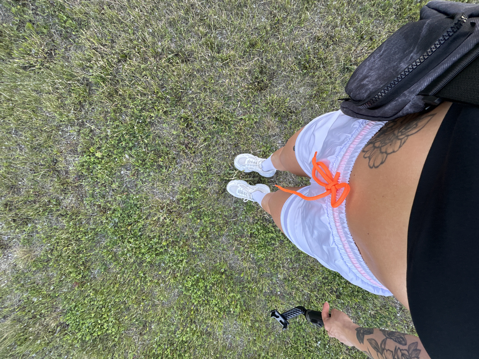 The image shows a top-down view of a person standing on a grassy area, likely a park or outdoor space in Yuzhnouralsk, Russia. The person is wearing white athletic shoes with white socks, white shorts with pink trim and an orange drawstring, and a black top. They are holding a black selfie stick in their left hand. A black fanny pack is worn across their body, resting on their hip. The ground is covered in green and brown grass with some patches of clover. The lighting suggests it is daytime.