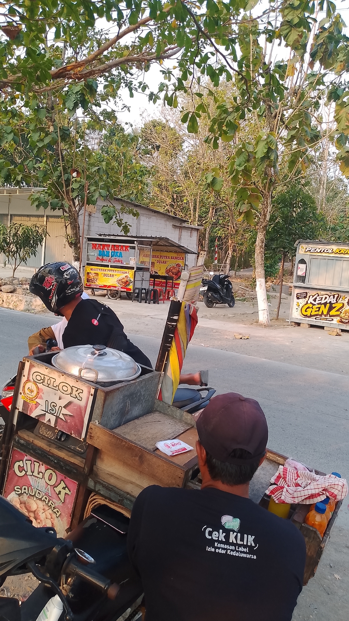 In Karanglangu, Indonesia, two men are seen near a roadside food stall. One man, wearing a black helmet with red and white accents, sits on a motorcycle. He wears a black long-sleeved shirt over a white shirt. His left arm is extended, potentially interacting with the motorcycle or a cart attached to it. The motorcycle has a metal cart on its rear, containing a large lidded pot and several boxes with labels that read "CILOK ISI" and "CILOK TIGA SAUDARA". A colorful striped flag is attached to the cart.
The second man, facing away from the camera, wears a dark baseball cap and a black t-shirt with white text that reads "'Cek Klik, Kemasan Label Izin edar Kedaluwarsa". He is positioned behind the motorcycle cart, facing the street. On the cart next to him are two bottles of orange liquid and a red and white checkered cloth.
In the background, a food stall with a white roof and grey walls is visible, displaying signs for "MARTABAK PUTRA BANGKA DULAH". Further down the street, another stall labeled "PENTOL KRI" and "KEDAI GENZ" is present. A black motorcycle is parked to the right of the "MARTABAK PUTRA BANGKA DULAH" stall. Trees with green and yellow leaves are abundant in the scene, suggesting a sunny day with sparse foliage on some trees, indicating a dry season. The ground is paved with asphalt and gravel.