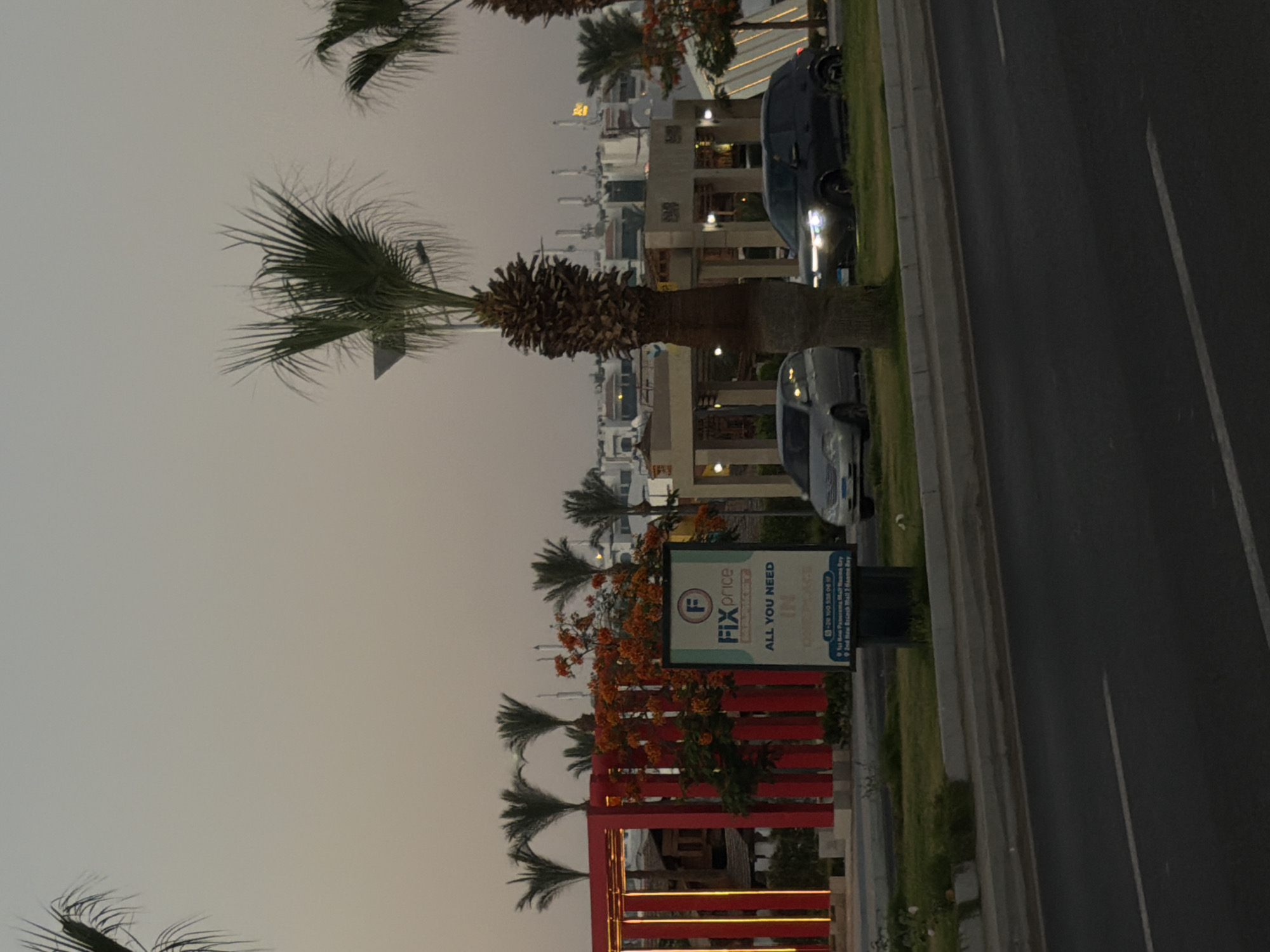 The image depicts a street scene in Sharm El Sheikh, Egypt, at dusk. The foreground shows a paved road with lane markings. A grassy median separates the road from a sidewalk area. On the sidewalk, several vehicles are parked: a silver sedan on the left and a dark-colored SUV on the right. Behind the vehicles, there is a red structure with vertical yellow lights, a billboard advertising "FIX PRICE," and several palm trees. In the background, buildings and more palm trees are visible against a hazy sky. The overall atmosphere is tranquil, typical of a suburban or commercial area at the end of the day.
