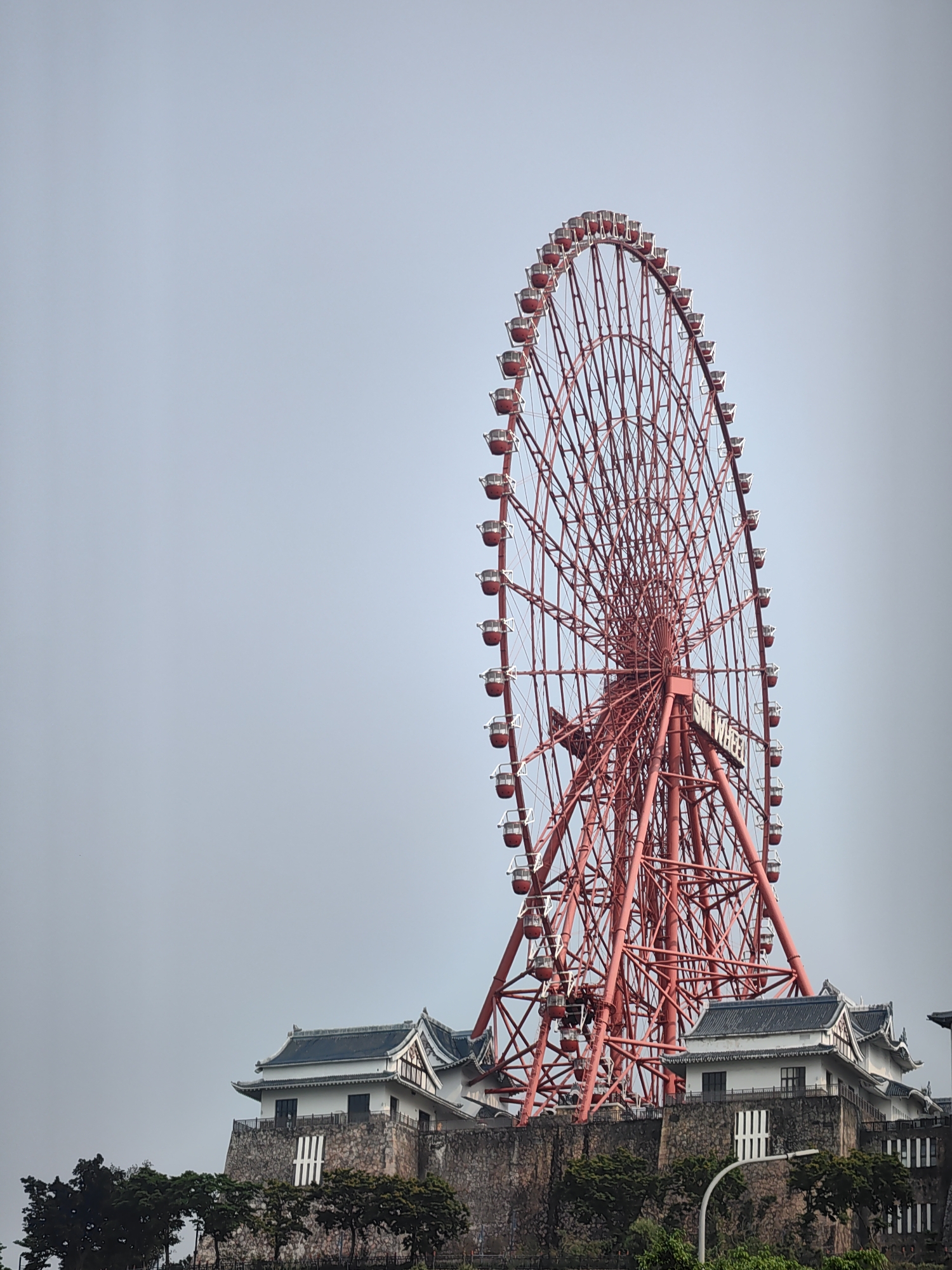 A red Ferris wheel stands tall behind a stone building with multiple white-painted structures and dark tile roofs. The building rests on a stone wall. Trees and greenery are visible in front of the wall. A light-colored sky provides the background. The Ferris wheel has passenger cars and the words "SUN WHEEL" on its support structure. The location is Hong Gai Ward, Vietnam.