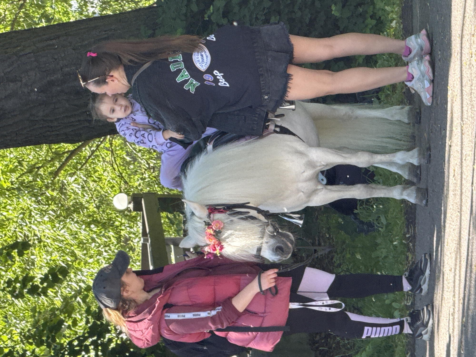 A young girl, dressed in a purple tracksuit with white lettering, sits atop a white pony adorned with a pink flower crown. She is held by a woman with long, dark hair pulled back in a ponytail, wearing a black denim jacket with patches and distressed denim shorts. To the left, another young woman wearing a pink puffer vest, black leggings with pink accents, and a dark baseball cap holds the pony's reins. They are standing on a paved path in an outdoor environment with lush green trees and foliage in the background. The scene appears to be a sunny day, possibly in a park or a recreational area. The location is not specified in the image or provided context, so it cannot be identified as Saint Petersburg, Russia.