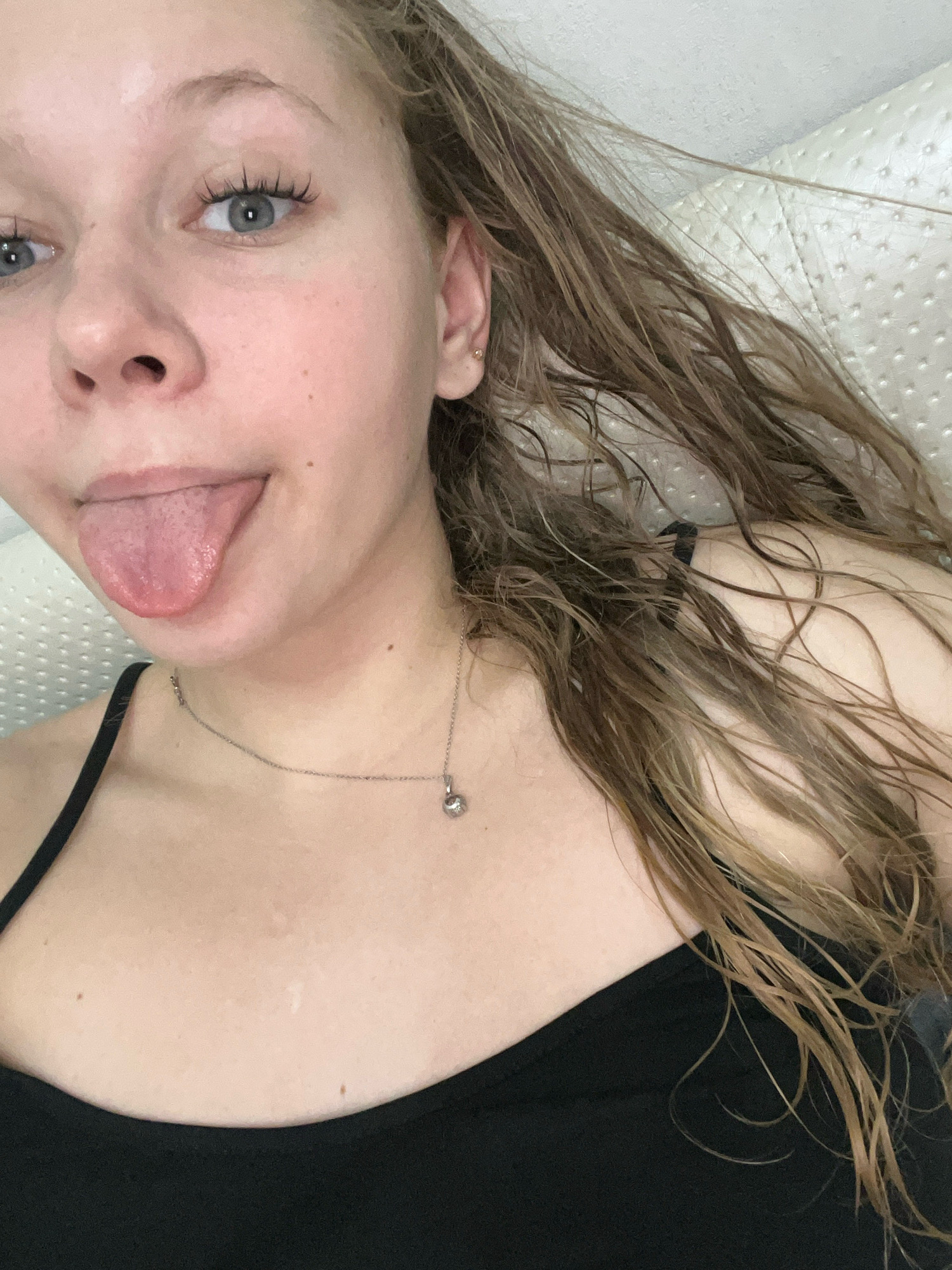 The image shows a close-up of a young woman with light-colored hair, which appears to be wet and is draped over her shoulder. She has blue eyes and is sticking out her tongue. She is wearing a black camisole with a thin strap, and a silver necklace with a small pendant. The background is a textured white surface, possibly a headboard or wall. There is no indication of the city of Perm, Russia, or any specific location.