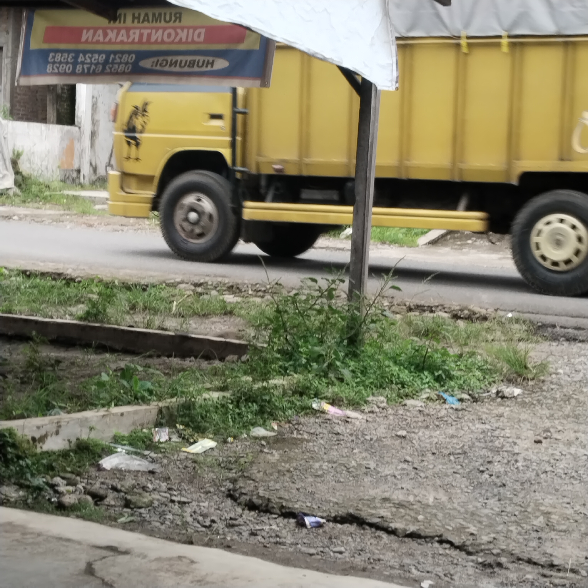 In the city of Talun Kenas, Indonesia, a yellow truck is parked on a road. The truck has a banner above it with Indonesian text. The ground around the truck is covered in gravel and patches of grass. There is also some debris scattered on the ground, including plastic bags and wrappers. The scene appears to be a typical roadside in a rural or semi-urban area.