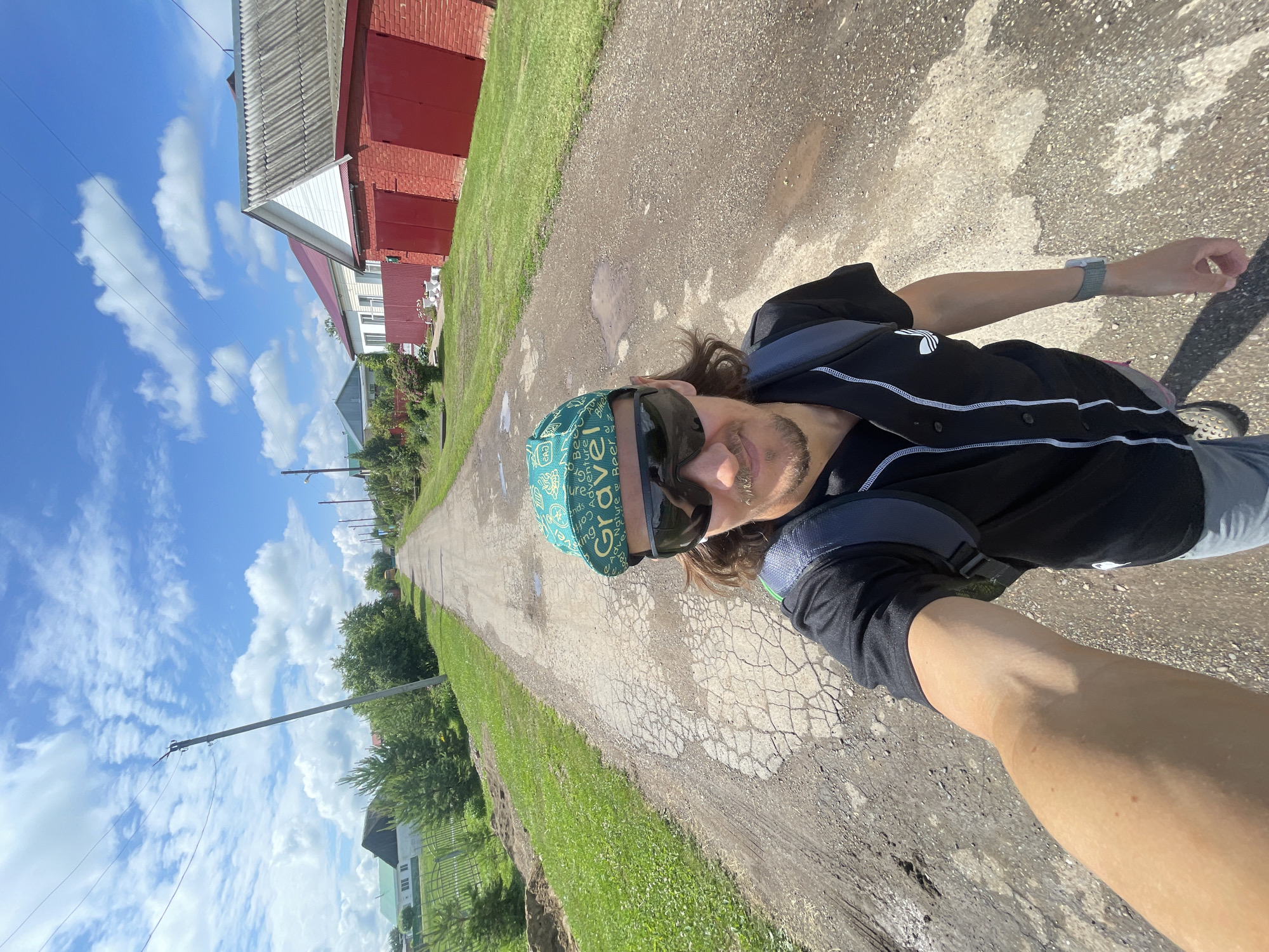 A man is taking a selfie while standing on a rural dirt road. He is wearing a black t-shirt with white piping, a green baseball cap with white lettering, sunglasses, and a grey backpack. The road is cracked and has several puddles. On either side of the road are grassy areas with trees and houses. The sky is blue with some white clouds.