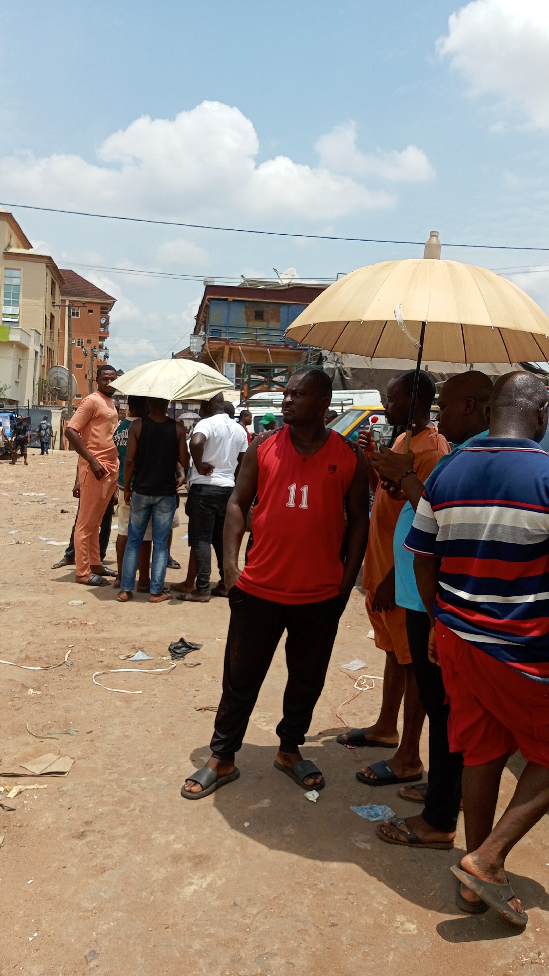 This image captures a bustling street scene in Ogbunike, Nigeria, under bright daylight. A group of approximately 10-15 men are gathered on an unpaved, dusty ground littered with debris. The sky above is clear with scattered white cumulus clouds.
In the foreground, a man in a red sleeveless shirt with the number "11" printed on it and black pants, wearing black slides, stands facing slightly to the right, looking into the distance. To his right, under a large, light-colored open umbrella, several men are congregated. One man wears an orange tunic and another a blue and red striped t-shirt. Another man under this umbrella appears to be holding a small red object. Further to the left, another large, light-colored umbrella provides shade for a smaller group of men, one wearing a black sleeveless shirt and blue jeans, and another in a white t-shirt. On the far left, a man in a peach-colored traditional outfit (tunic and trousers) stands, also wearing sandals.
The men appear to be casually interacting, standing, and observing their surroundings. Their postures suggest a relaxed gathering or waiting. Partially visible vehicles, including what looks like a white bus or van and a yellow commercial vehicle, are in the background. The background also features multi-story buildings, some with satellite dishes, indicating an urban or semi-urban environment. The strong shadows suggest it is midday or early afternoon, requiring the use of umbrellas for sun protection. The overall atmosphere is casual and typical of a local street gathering.