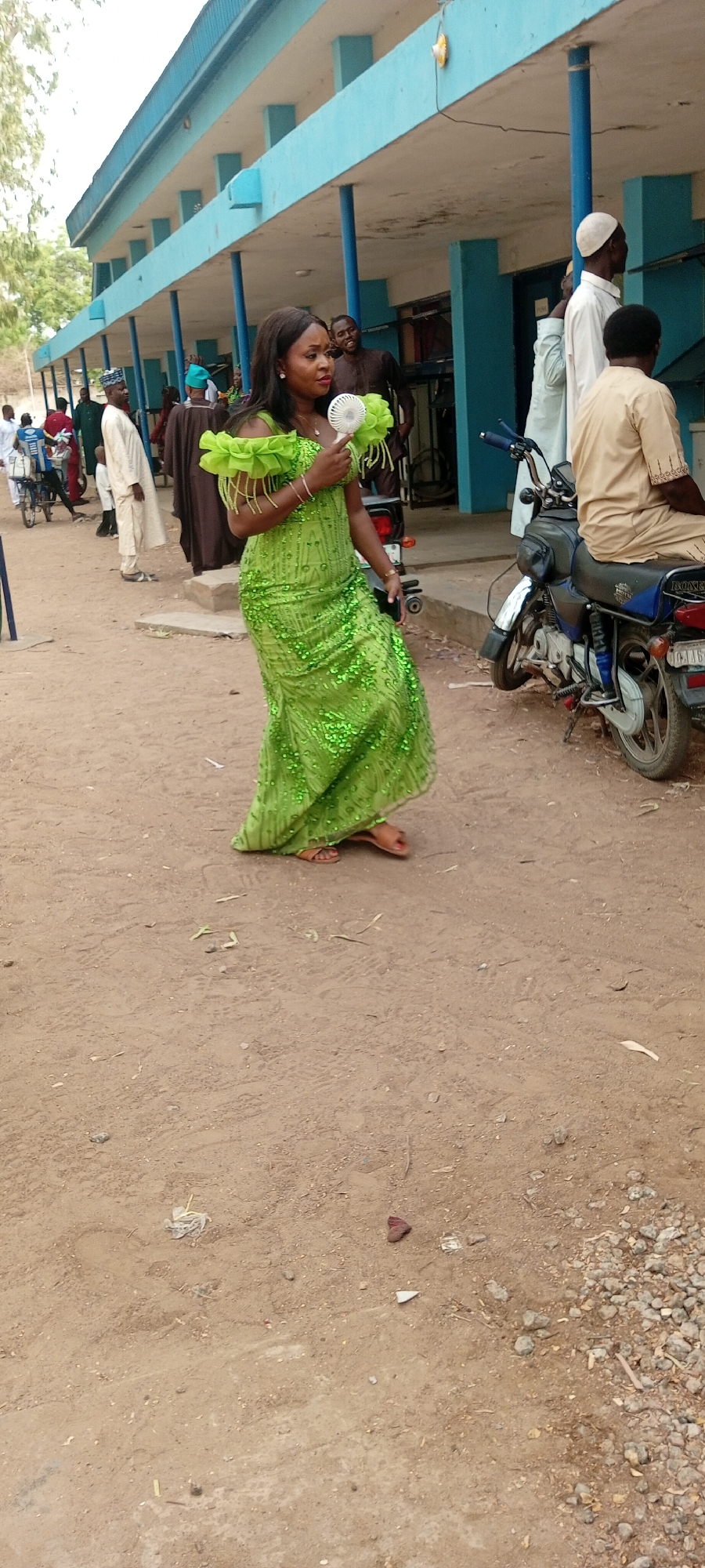 The image, taken in Fagge D2, Nigeria, shows a woman in a bright green, floor-length dress with ruffled shoulders walking on a dirt surface. She is holding a small, white, handheld fan. In the background, a row of buildings with blue trim and support columns lines the street. Several people are standing around, some in traditional clothing. A parked motorcycle is visible near the building. The ground appears to be a mix of dirt and gravel, with some small pieces of debris scattered around.