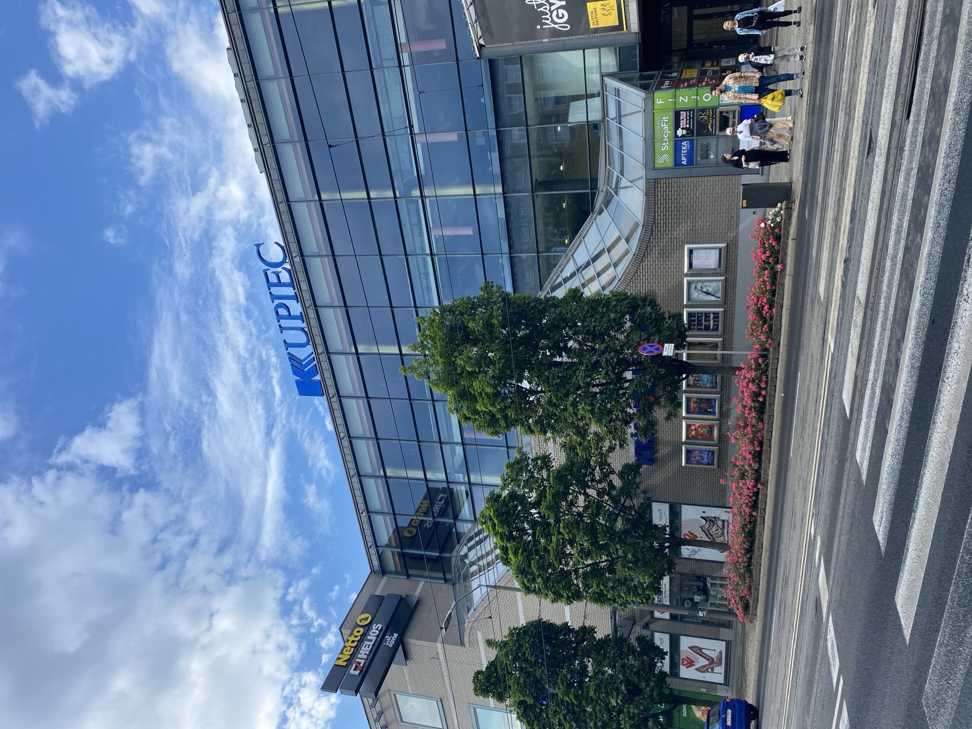 The image shows a street scene in Szczecin, Poland, featuring the Kupiec shopping center. The building has a modern design with a large glass facade. Above the entrance, a blue sign reads "KUPIEC". On the left side of the building, there are signs for "Netto" and "HELIOS". In front of the building, there is a flower bed with pink flowers. Several people are standing on the sidewalk, waiting to cross the street at a crosswalk. A blue car is parked on the left side of the street. The sky is cloudy.