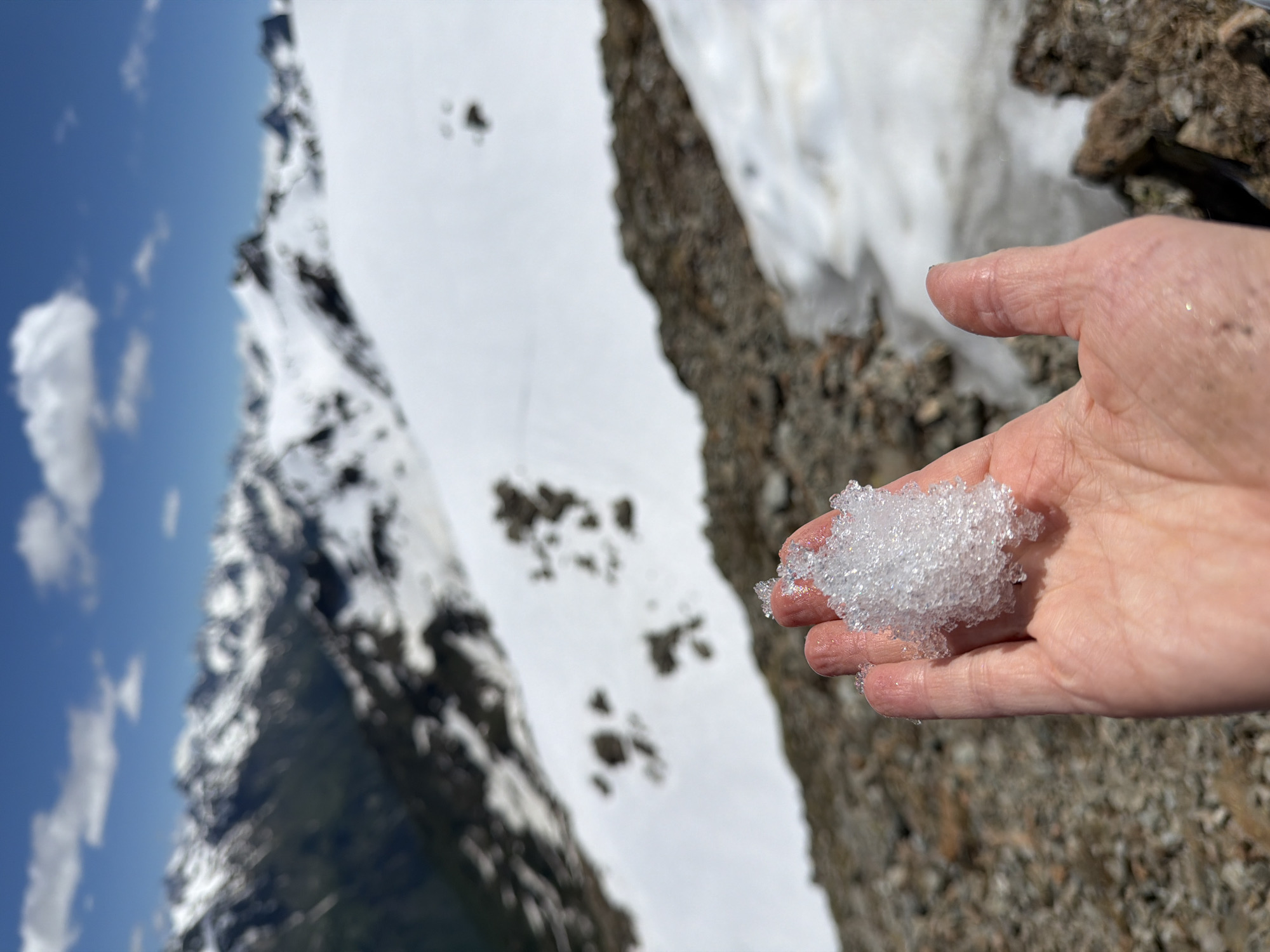 A close-up shot shows a person's hand holding a clump of snow. The hand is lightly tanned with some visible veins. The snow is white and crystalline, appearing to be melting slightly. In the background, a snow-covered mountain slope is visible, with patches of dark rock. Above the mountain, a bright blue sky with fluffy white clouds suggests a sunny day. The overall scene conveys a sense of being in a cold, mountainous environment, possibly during a hike or an outing in nature. The lighting suggests it is daytime, likely late morning or afternoon due to the brightness of the sky. The location is identified as Верхне-Тебердинское сельское поселение, Russia.