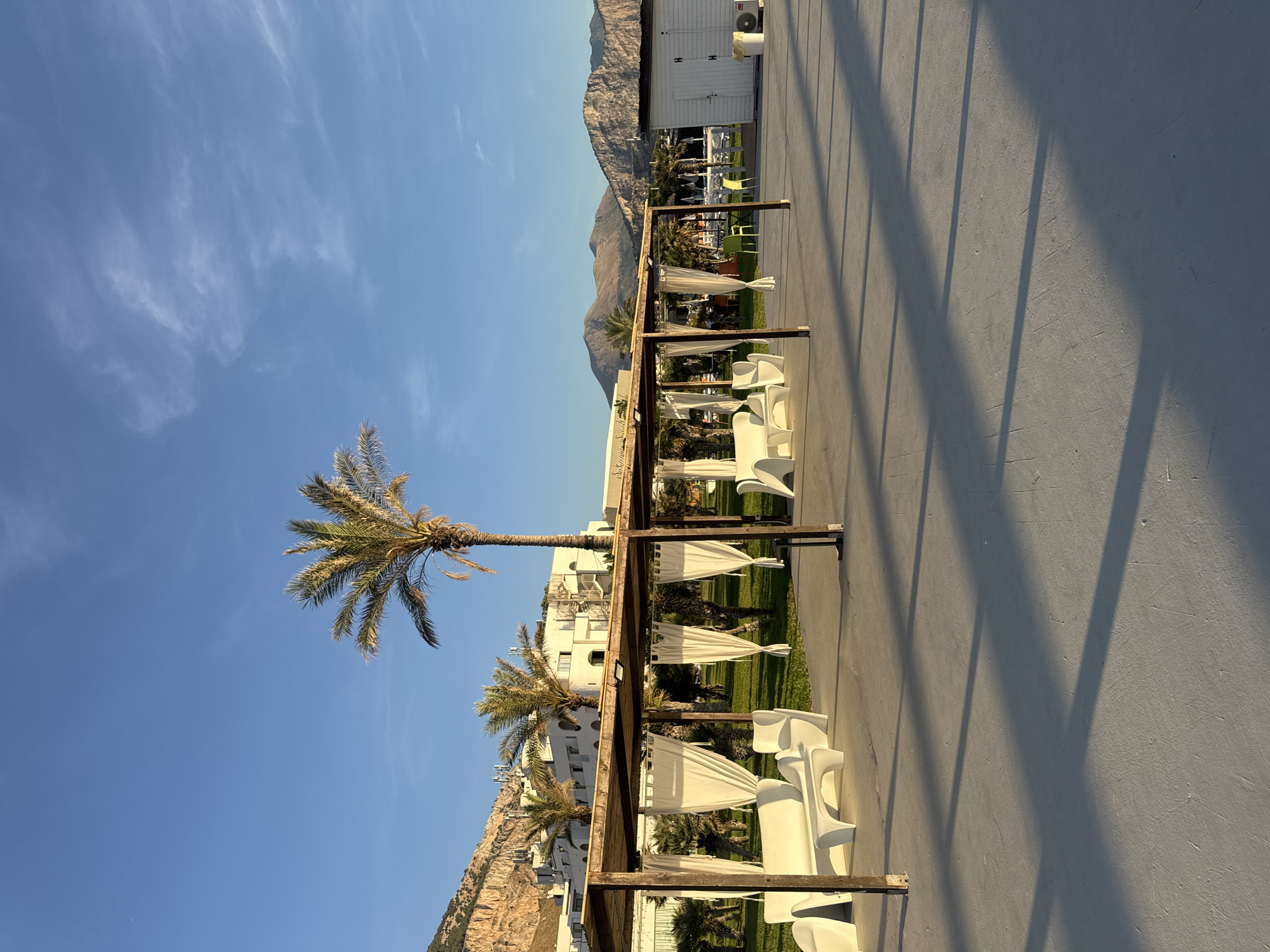 The image depicts a serene outdoor seating area with a wooden pergola, white sofas, and white curtains, set against a backdrop of palm trees and mountains. The location is identified as Isola delle Femmine, Italy. The scene appears to be a resort or beach club, with a clean, modern aesthetic. The sun is low in the sky, casting long shadows across the paved ground. In the background, a white building with arched windows is visible, along with more palm trees and the rugged terrain of the mountains. The sky is a clear blue with a few wispy clouds. There are no people visible in the image.