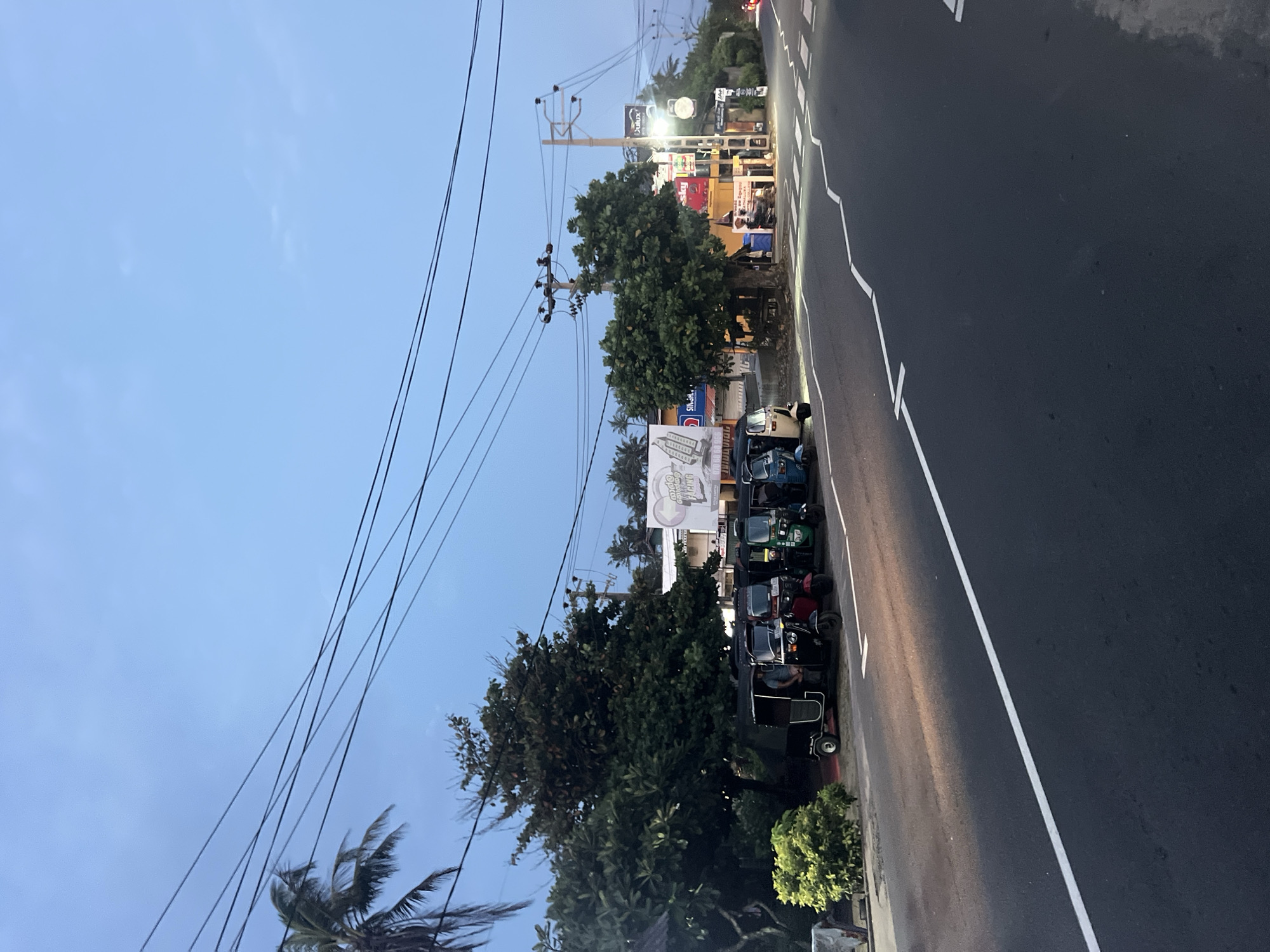 The image shows a street scene in Kathaluwa, Sri Lanka. The street is paved with asphalt and has white lane markings. Several three-wheeled vehicles are parked along the side of the road. There are buildings and trees visible in the background, including a billboard. Overhead power lines stretch across the sky. The sky is light blue, suggesting it is daytime or early evening. The scene appears calm and ordinary.