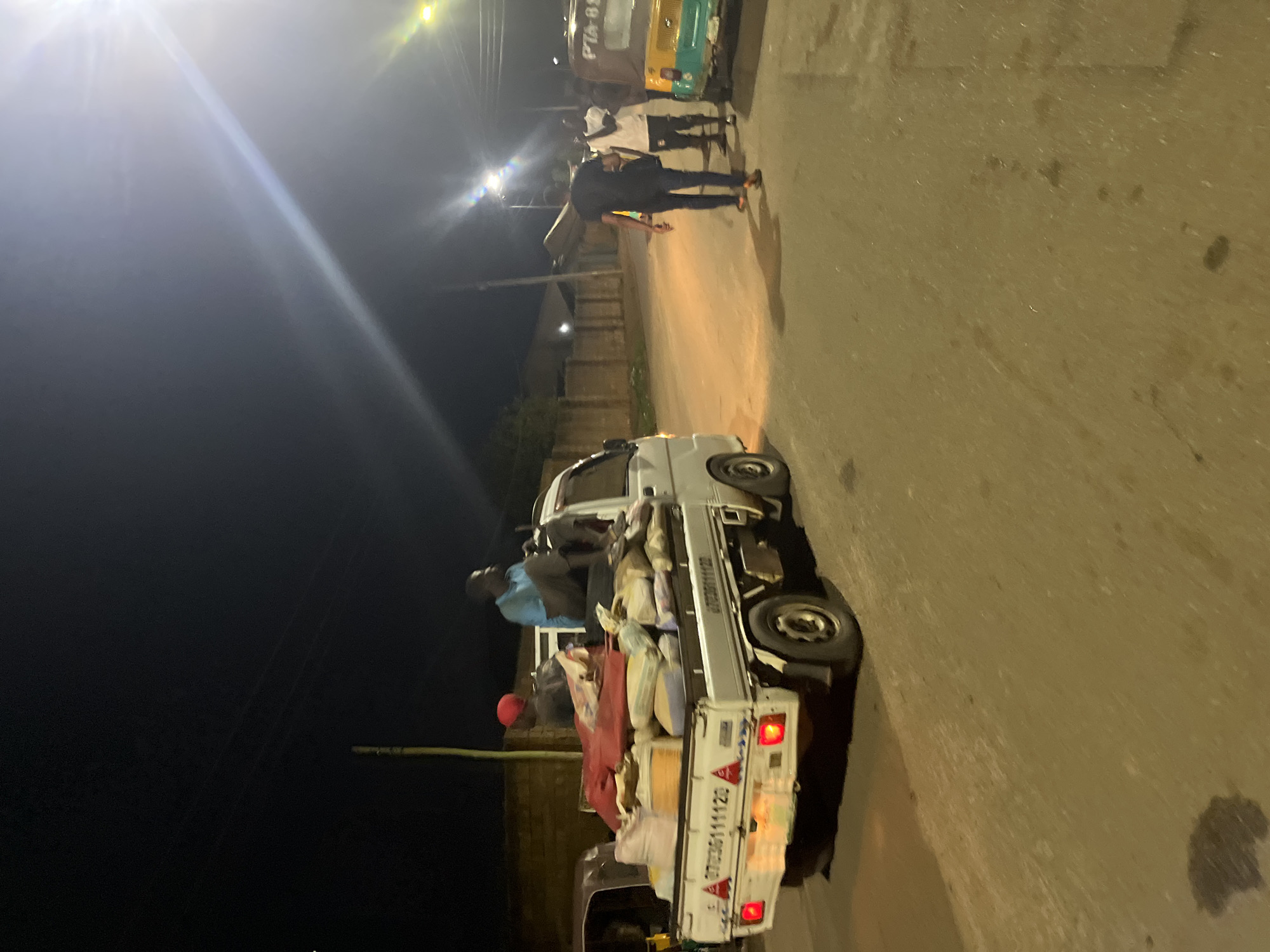 The image captures a scene at night in Jos, Nigeria. A white truck is parked on a paved road, its flatbed loaded with sacks and covered with a red tarp. At least three people are seated on top of the load, while others stand on the road nearby. The lighting is provided by streetlights, casting bright spots and long shadows. In the background, a vehicle resembling a taxi or bus is parked, and additional people are visible. The area seems to be a residential or commercial district.