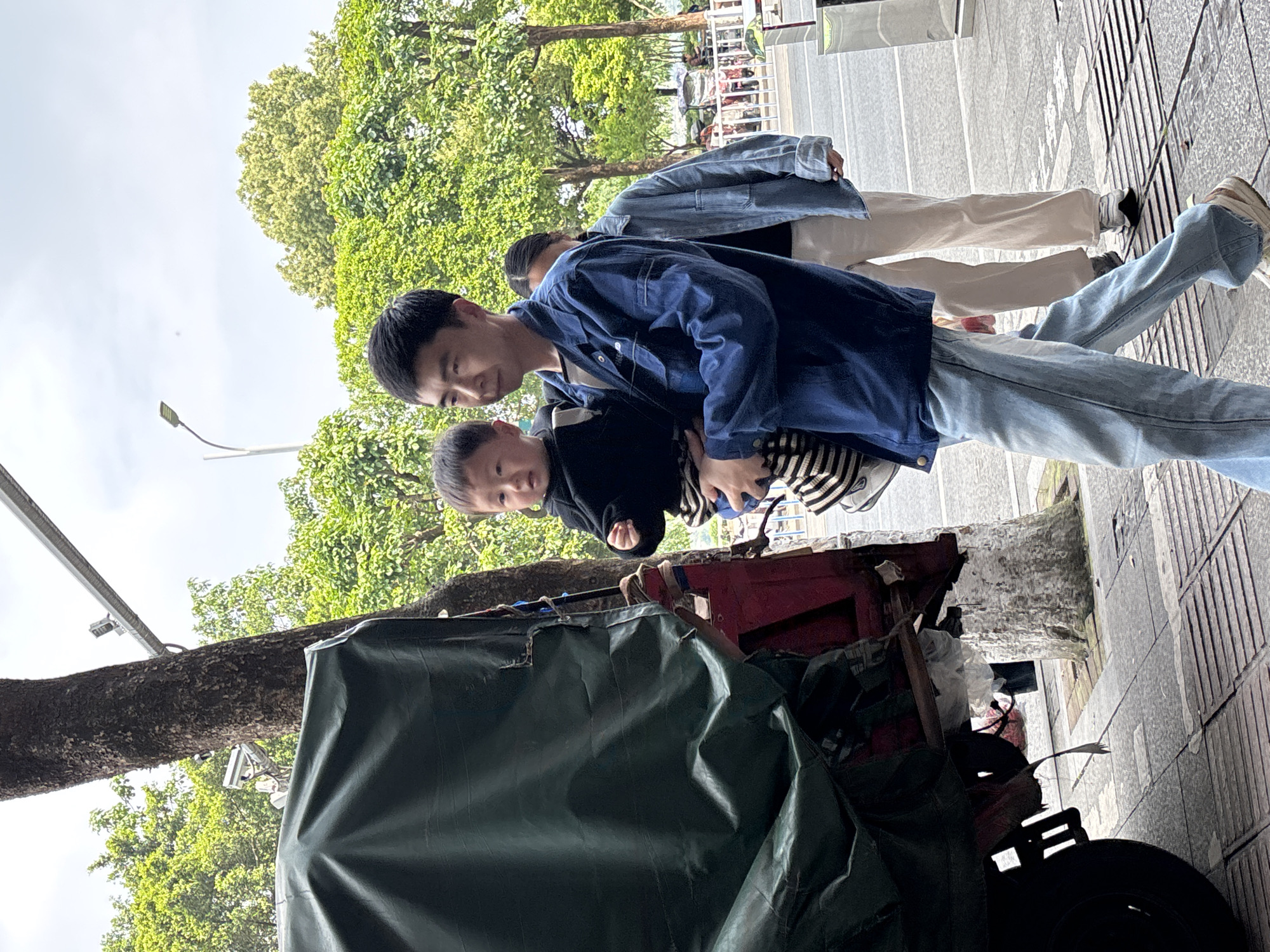 The photograph depicts a man carrying a young child, likely a toddler, on a sidewalk adjacent to a city street. The man is wearing a blue denim jacket and light-wash jeans. The child is wearing a dark jacket over a striped shirt. Another individual, wearing a denim jacket and light-colored pants, is walking behind the man. The setting appears to be an urban environment, as evidenced by the sidewalk, street, trees, and building in the background. A covered cart or stall is visible in the foreground, suggesting a possible commercial street vending operation. The photo was taken in Jiangjin, China.