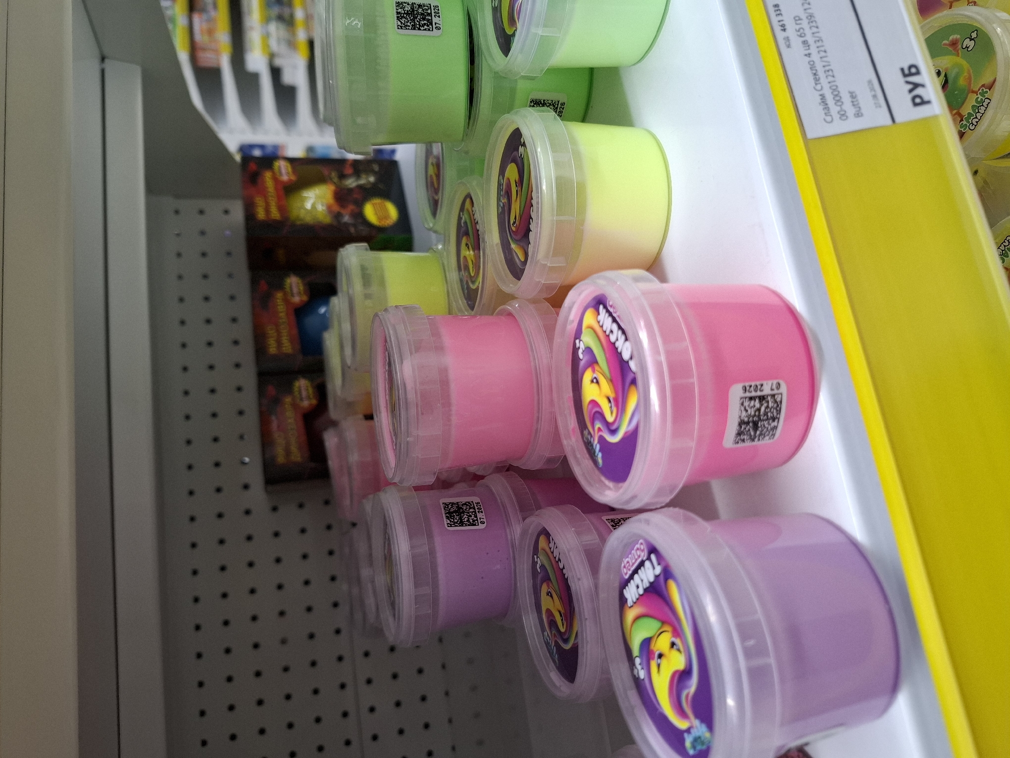The image shows a retail shelf in Tyumen, Russia, displaying various colorful tubs of slime. The slime is presented in clear plastic containers with colorful lids, and the contents themselves are vibrant shades of purple, pink, and yellow. Some of the tubs have labels with barcodes and text, including what appears to be product information and expiry dates. In the background, there are boxes with imagery that suggests they contain toys, possibly dinosaur-themed. The shelf is white, with a yellow price tag or shelf edge strip visible in the foreground. The lighting is bright, typical of a store environment. No people are visible in the image.