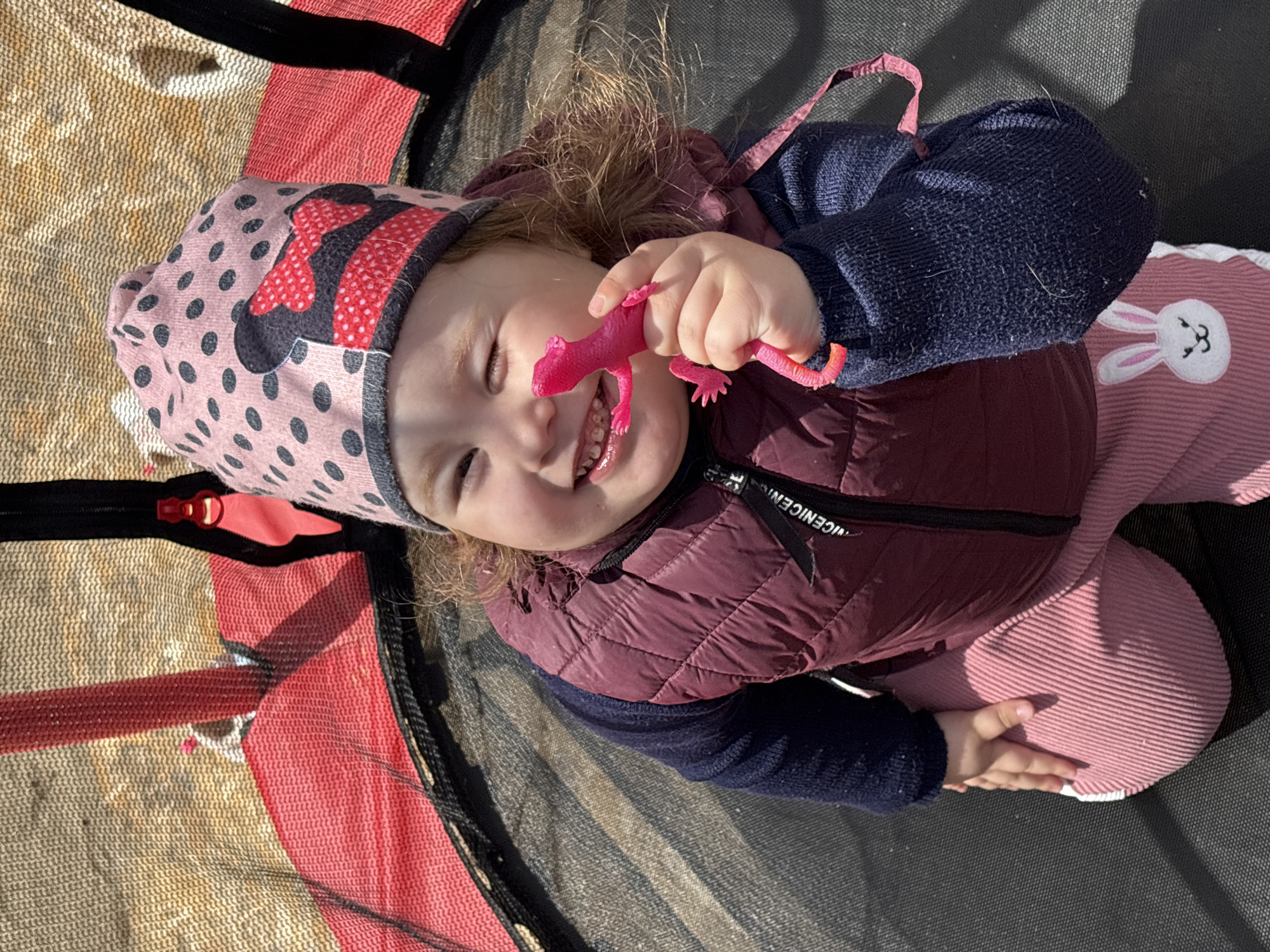 A young child, likely a toddler, is lying on a trampoline. The child is wearing a pink hat with a Minnie Mouse design, a navy blue long-sleeved shirt, a maroon puffer vest, and pink corduroy pants with a white bunny appliqué. The child is smiling and holding a pink toy lizard in their hand. The trampoline has a red and yellow safety pad around the edges. The location is unknown and there is no evidence that it is in the city of Prikamskii, Russia.
