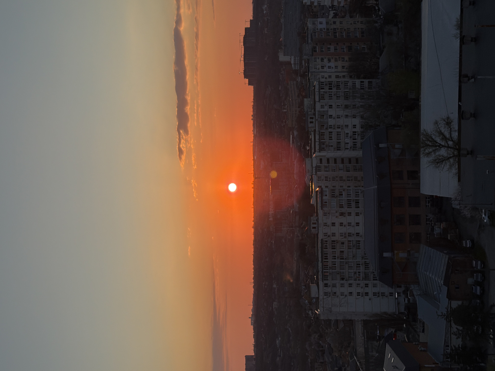 The image shows a sunset over a city. The sun is low on the horizon, casting an orange glow over the sky and the buildings below. In the foreground are several multi-story apartment buildings. The image is likely from Azov, Russia, based on the urban landscape and architectural style.