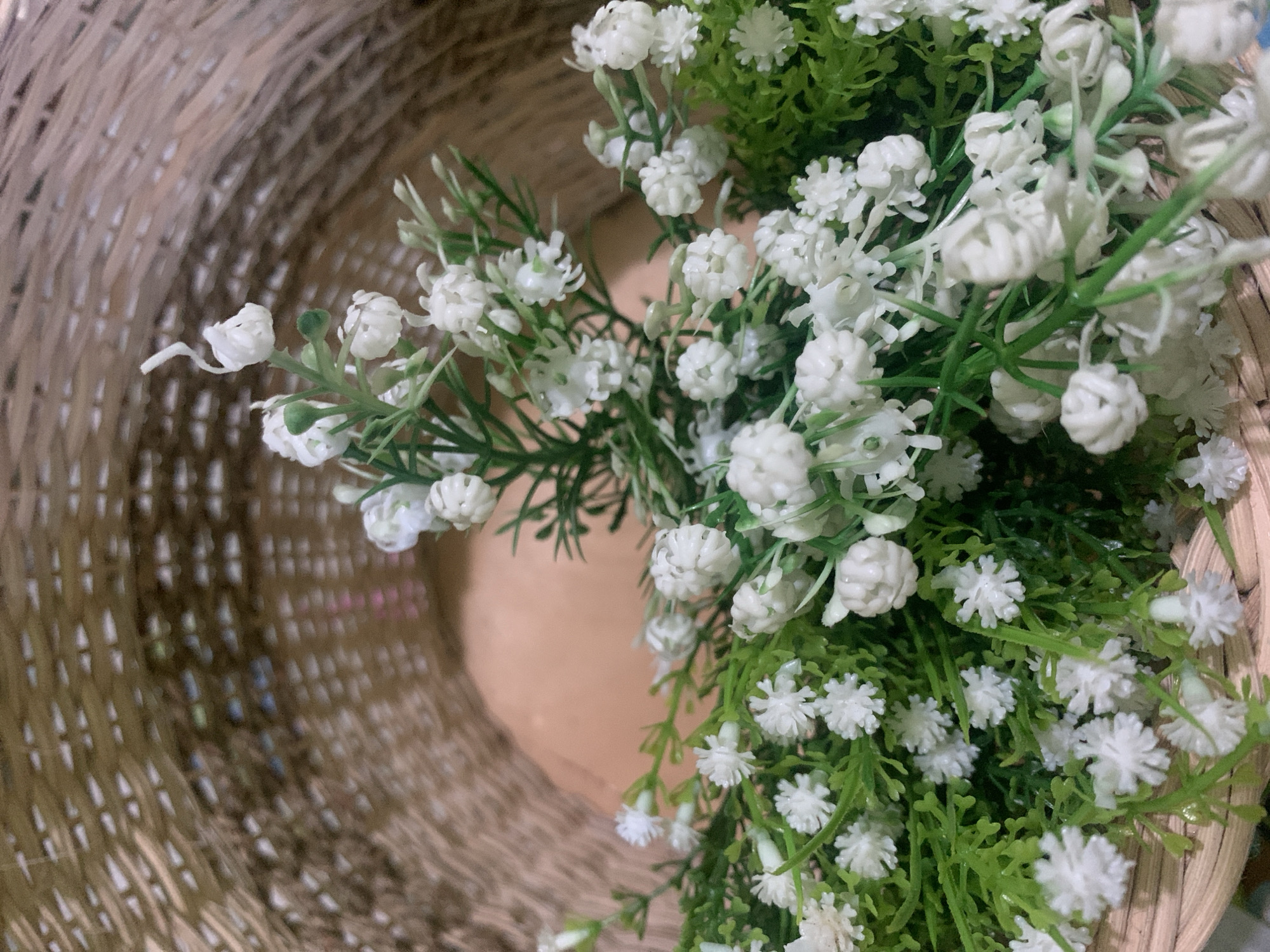 The image shows a close-up view of white artificial flowers with green stems and leaves inside a woven basket. The basket is made of light brown straw-like material and has a textured, layered appearance. The flowers are small and delicate, with some clustered together and others appearing individually. The background is blurred, suggesting a shallow depth of field. There are no people or specific landmarks visible in the image. The scene is indoors or in a protected outdoor area. The lighting appears natural and soft. The location is not specified in the image.