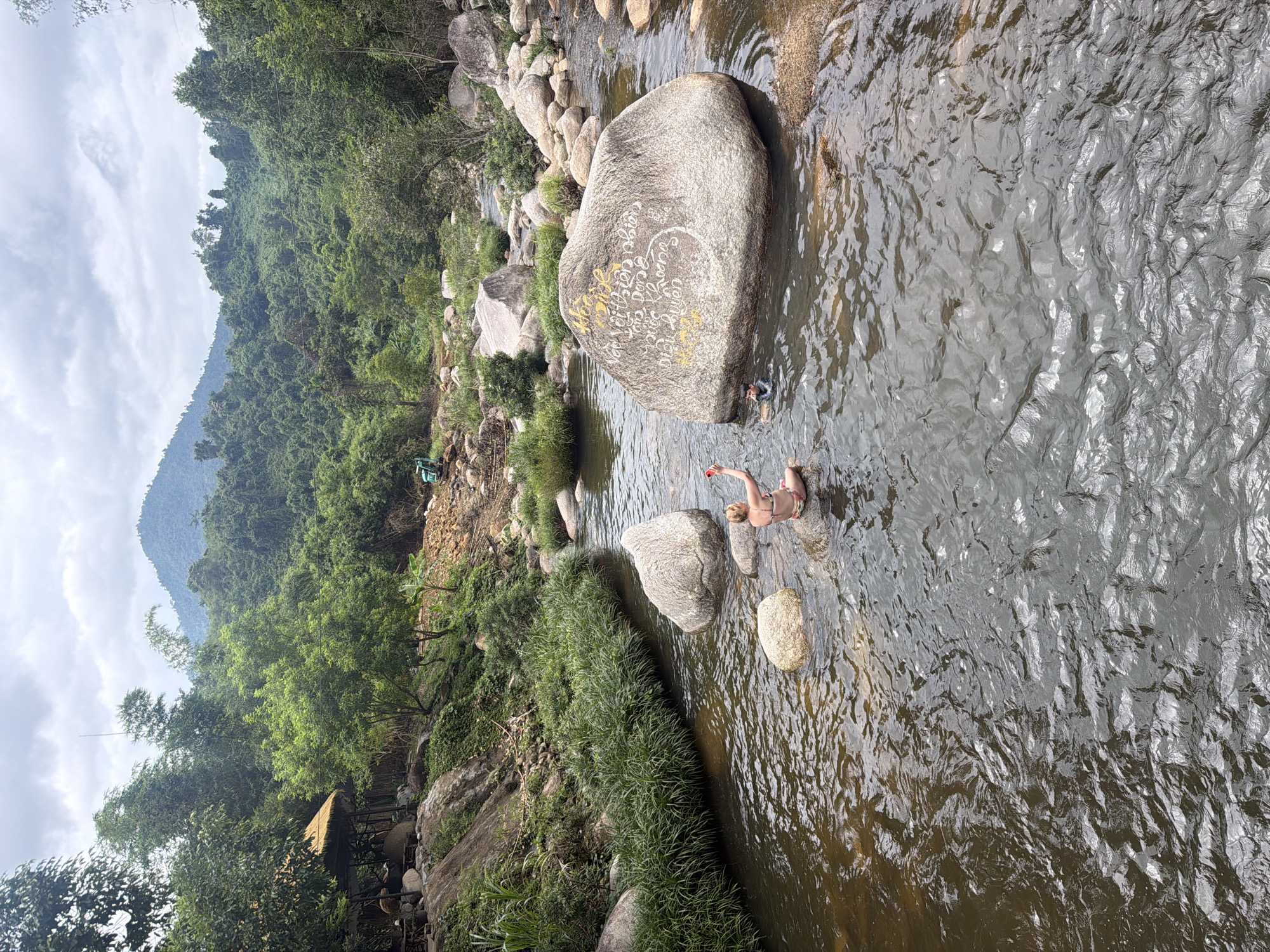 A wide shot captures a serene natural landscape with a shallow, rocky river flowing through it. On the left bank, a rustic thatched-roof structure with wooden posts and seating is nestled amongst lush green vegetation. Further back, a construction vehicle is visible on a dirt path. The river is populated by large, smooth boulders, some of which are submerged. In the middle of the river, a person wearing a bikini sits on a rock, holding a red object, possibly a phone. Nearby, a smaller figure, possibly a child, is partially submerged in the water. A large boulder in the river bears inscriptions in yellow and white paint. The background is dominated by verdant, tree-covered mountains under a cloudy sky. The scene is located in Khanh Vinh, Vietnam.