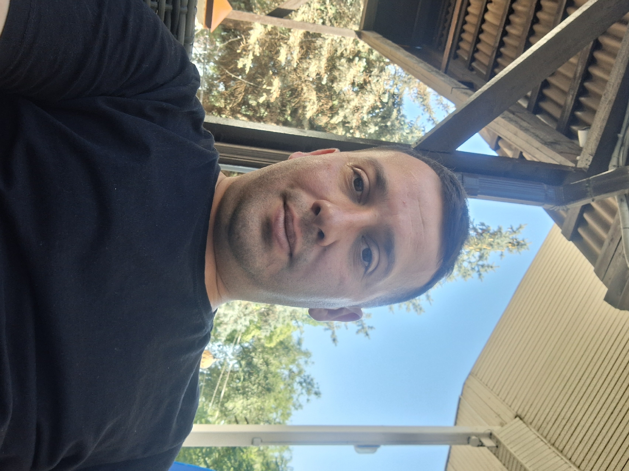 A man in his late 30s or early 40s, with fair skin, short dark hair, and a clean-shaven face, is seated outdoors, likely on a patio or under a covered structure. He is wearing a black t-shirt and is looking directly at the camera. The background shows trees and a clear blue sky, suggesting a sunny day. Part of a wooden structure with a slatted roof is visible above and to the left of the man, and a light-colored ceiling is visible to the right. The overall setting appears to be casual and relaxed. The provided image and its crops do not contain any information that would allow for determining the specific location in Rostov-on-Don, Russia, or any other geographical details.
