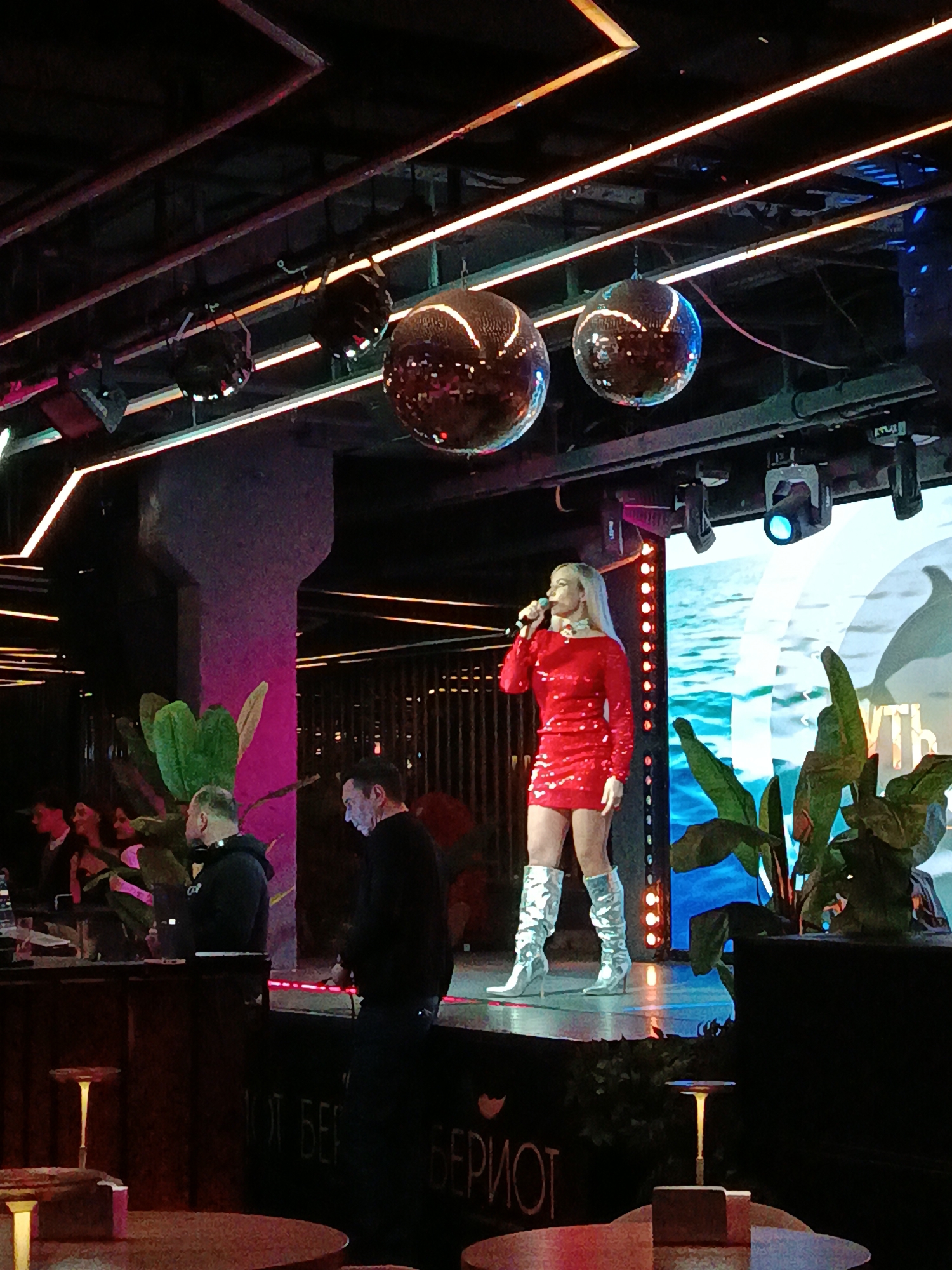 The image captures a scene on a stage in an indoor venue, potentially a nightclub or restaurant, located in an unknown city. A woman with blonde hair is the central figure, standing on the stage and singing into a microphone. She is wearing a red sequined dress and silver knee-high boots. Behind her, there's a large screen displaying a water scene with dolphins.
Above the stage, disco balls are suspended, reflecting light. Stage lighting fixtures are also visible.
In the foreground, there are tables, indicating the audience area. Several people are present, including a man in a black hoodie standing near the stage and several seated at a table. Some greenery decorates the stage and possibly the surrounding area. The text "ОТ БЕЕ" is visible on the stage front. The environment suggests a live performance setting with a focus on entertainment and possibly dining.
