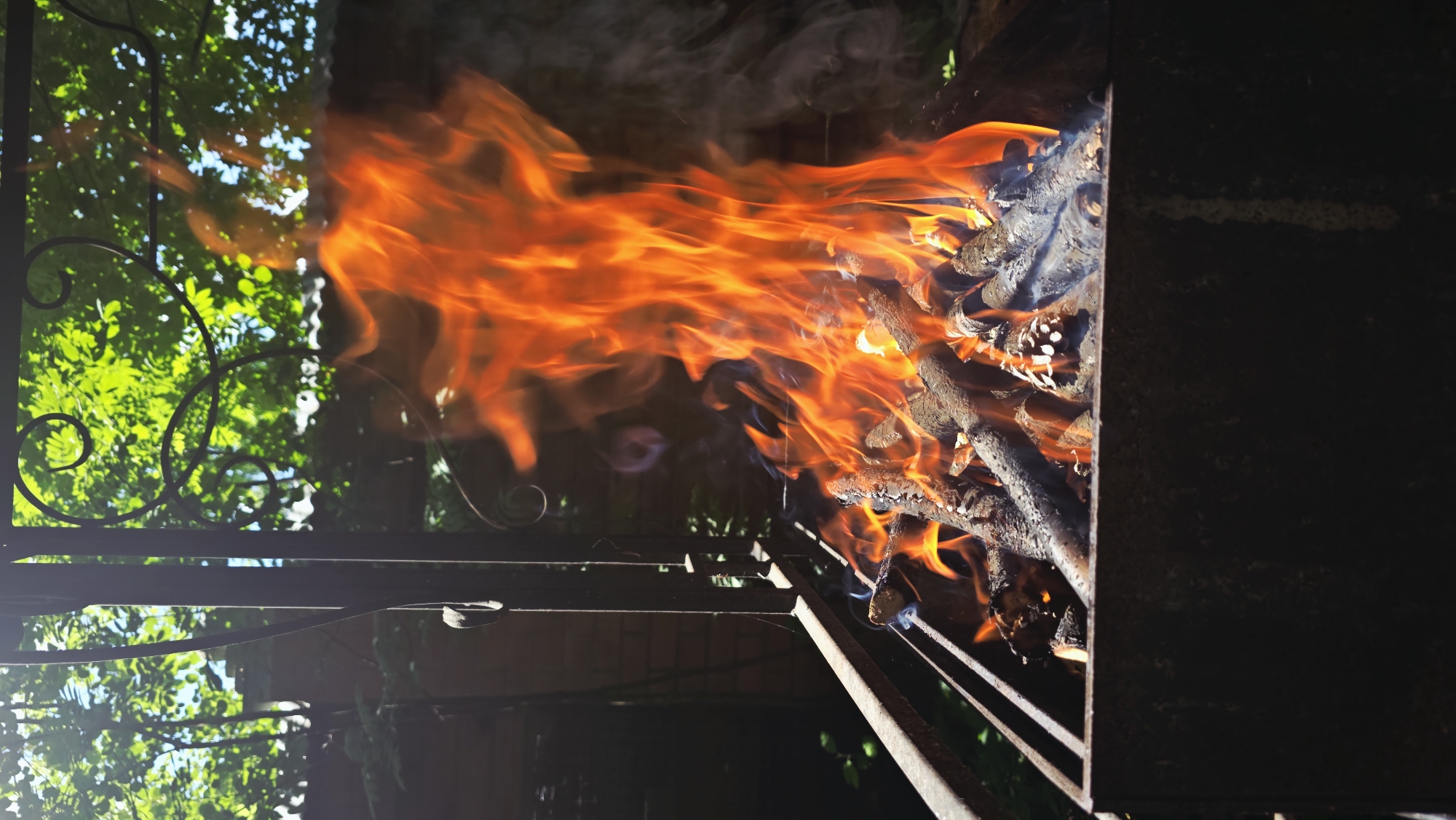 A close-up, low-angle outdoor shot captures a rustic barbecue grill ablaze with vibrant orange flames. The grill, constructed of dark metal, is filled with burning wood, emitting wisps of smoke that curl upwards. The fire consumes several logs and a pine cone, creating a dynamic and energetic display. In the background, lush green foliage and a clear blue sky suggest a warm, sunny day. The scene is framed by dark metal bars, possibly part of the grill's structure or a nearby fence, with decorative scrollwork visible at the top. The overall impression is one of a lively outdoor gathering, possibly in a backyard or park setting within Il'inskiy, Russia. There are no people visible in the frame.