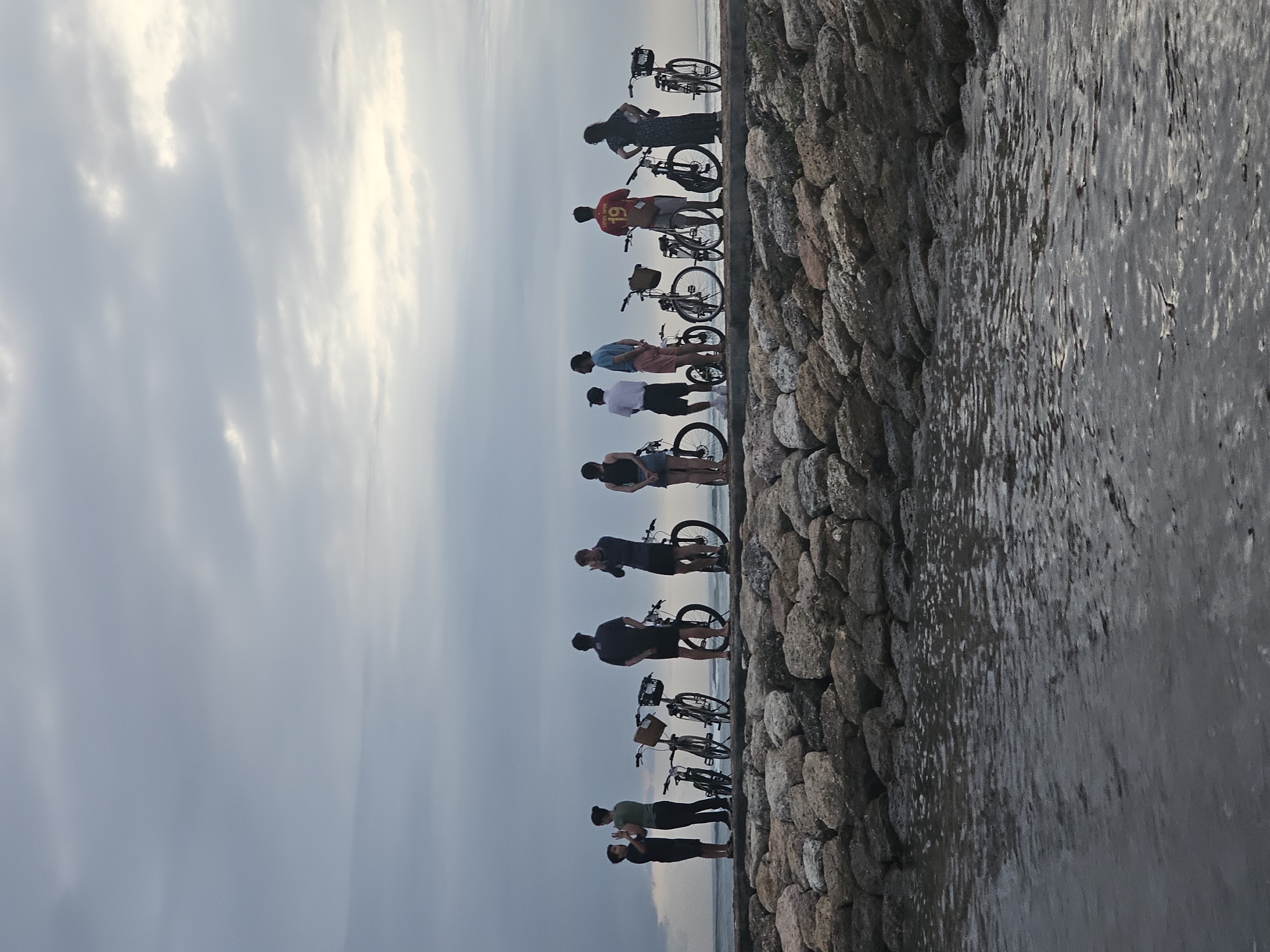 In Denpasar, Indonesia, a group of nine individuals stands with their bicycles along a stone wall that appears to be a breakwater or sea defense structure. The wall separates a wet, dark gray surface, possibly a walkway or the receding tide, from the sky. The sky is overcast with gray clouds. The individuals are oriented in the same direction, away from the camera and towards the horizon. They are dressed in casual clothing, with one person wearing a red jersey with the number '19' on the back. The bicycles are a mix of models with and without baskets. The scene suggests a casual outing or a break during a bicycle ride along the coast.