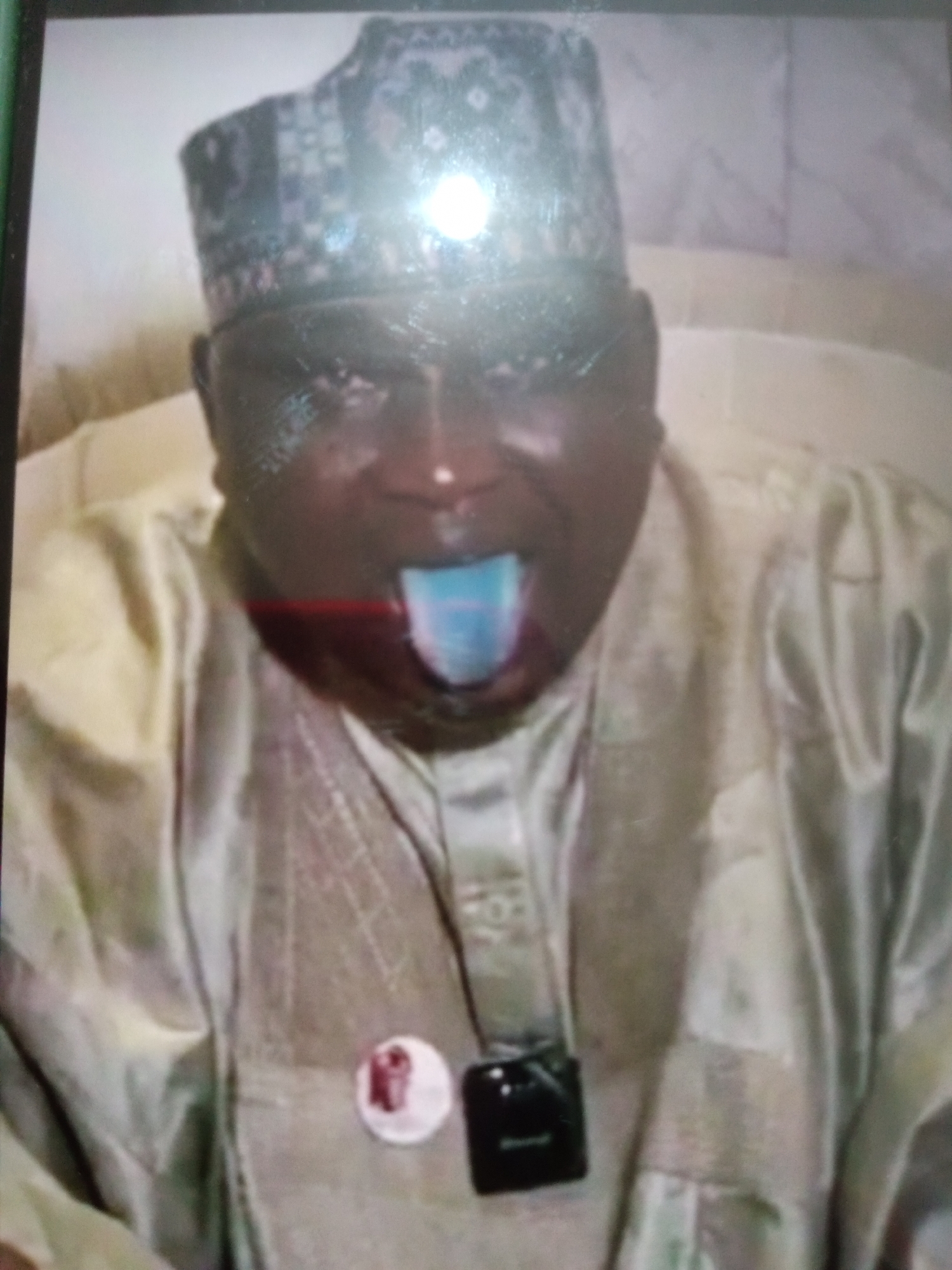 A close-up, slightly blurry, medium shot shows a dark-skinned man wearing a traditional cap and a light-colored, possibly silk, robe. The man is seated indoors, possibly in a residential setting in Jalingo, Nigeria. His tongue, which is blue, is sticking out. A small, circular pin with an image on it is visible on his robe, just below his neck. A small black device, possibly a microphone or a pager, is also attached to his robe. The background is indistinct and appears to be a wall or drape, suggesting an indoor environment. The lighting is somewhat uneven, with a bright glare on the man's cap.