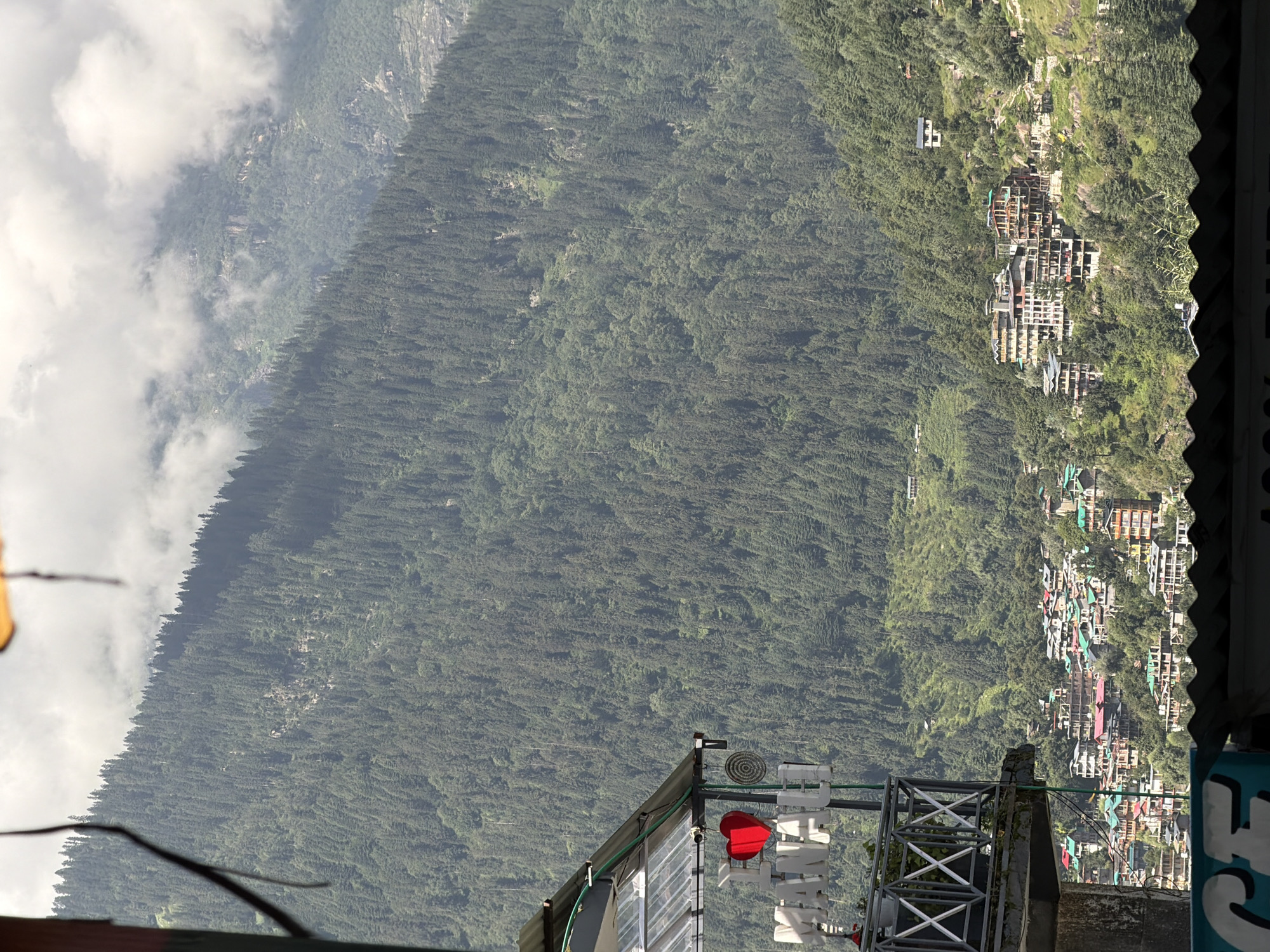 The image shows a view of the town of Vashisht, India, nestled in a valley surrounded by lush green mountains. In the foreground, a portion of a building with a metal roof and a balcony is visible. On the balcony, a large red heart-shaped object is mounted next to white letters that spell "MANALI". The town of Vashisht is characterized by its densely packed buildings with colorful roofs, constructed on the slopes of the mountains. The mountains themselves are covered in a thick forest of evergreen trees, with some patches of rocky terrain. The sky above is partly cloudy.